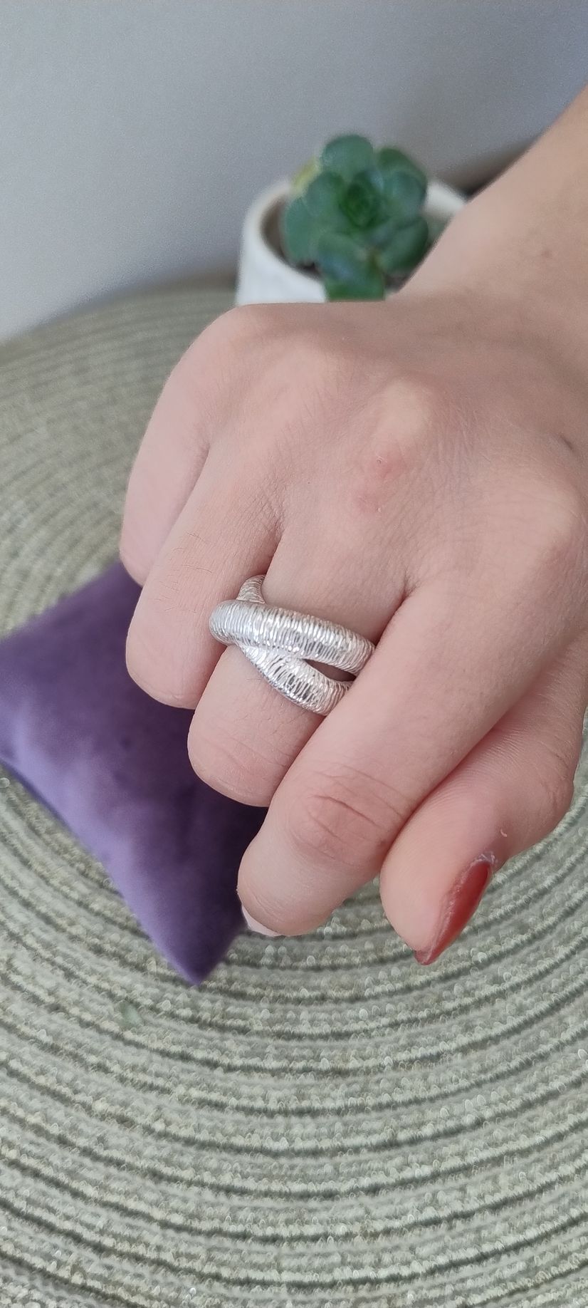Una mano con un anillo plateado cruzado descansa sobre un cojín morado. Detrás de la mano hay una pequeña planta suculenta.