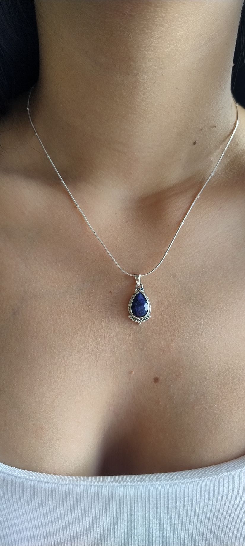 Primer plano de un collar de plata con un colgante azul en forma de lágrima en el cuello y el pecho de una persona.