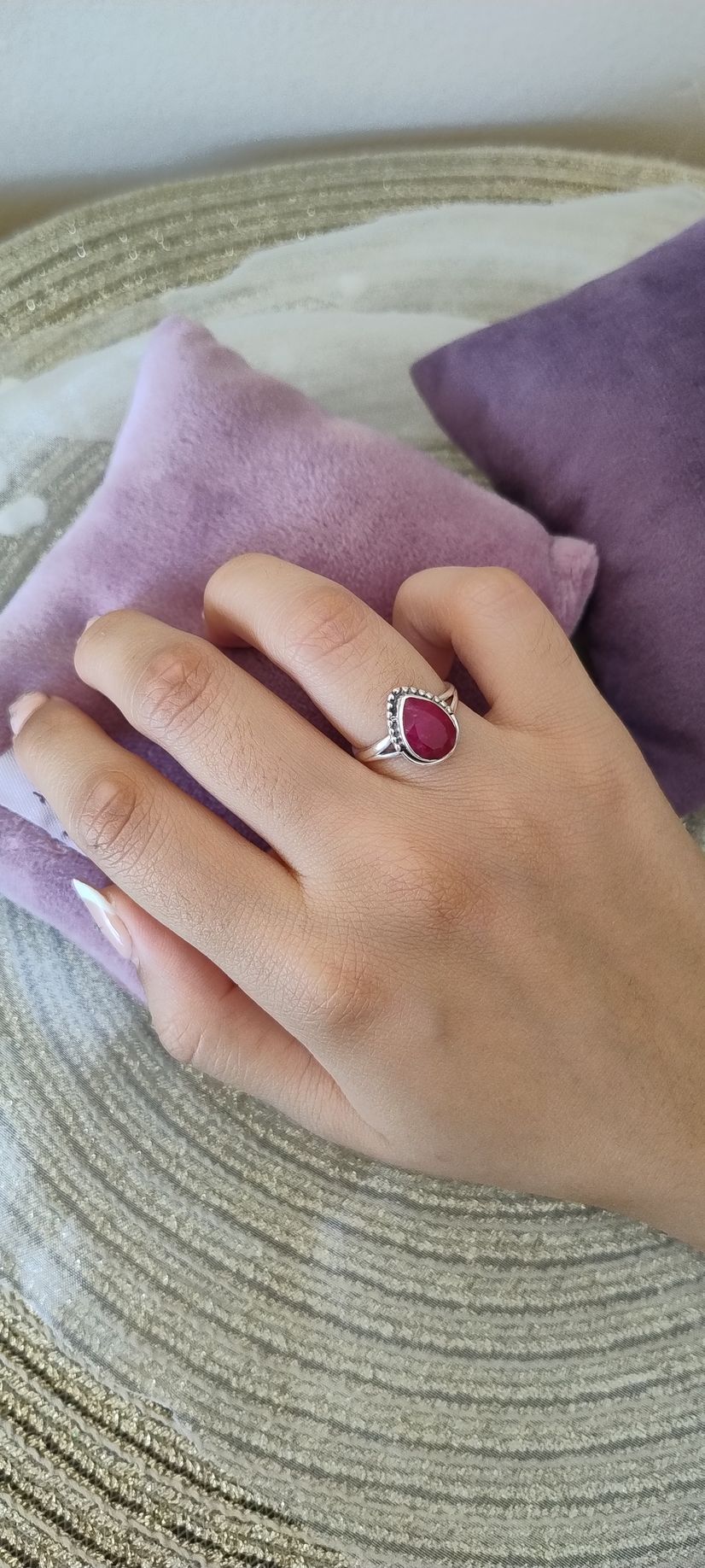 Una mano que lleva un anillo de rubí descansa sobre almohadas moradas sobre una superficie tejida de color beige.