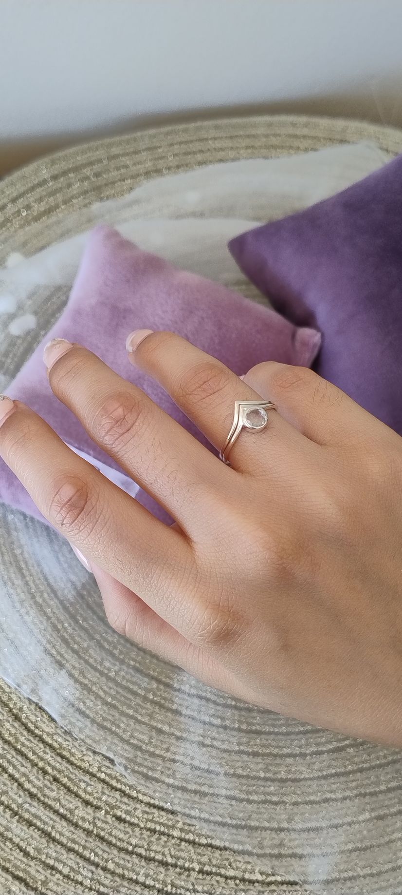 Una mano con un anillo de diamantes descansa sobre cojines morados, encima de un sombrero de mimbre.