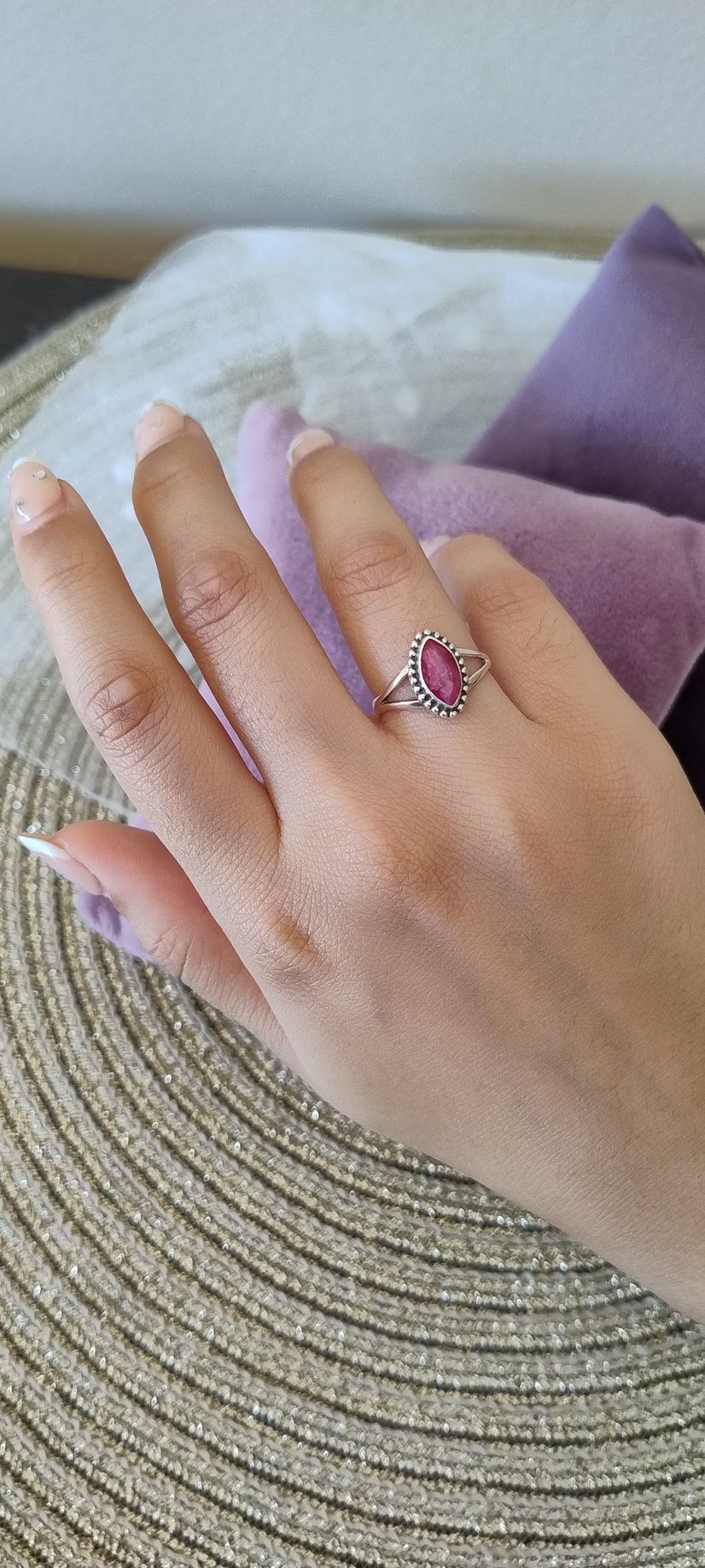 Una mano con un anillo de piedra preciosa rosa descansa sobre un sombrero de paja tejido, con una almohada morada al fondo.