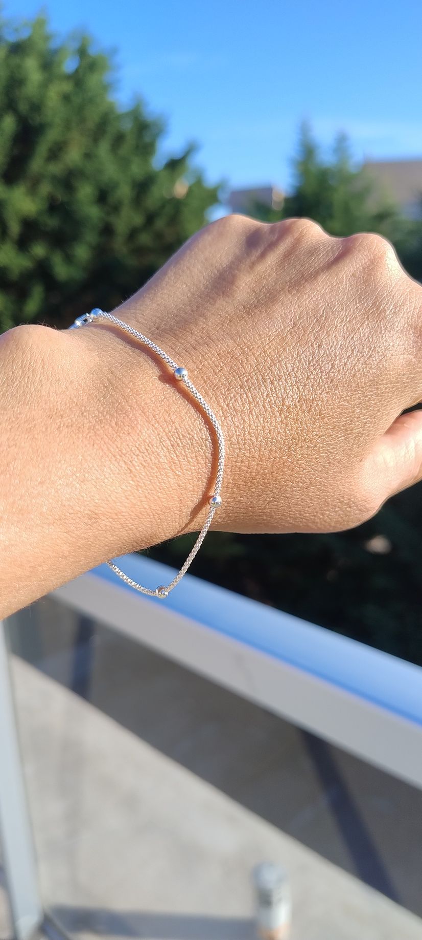 Mano con una pulsera de plata bajo la luz del sol, con árboles y la barandilla de un balcón al fondo.