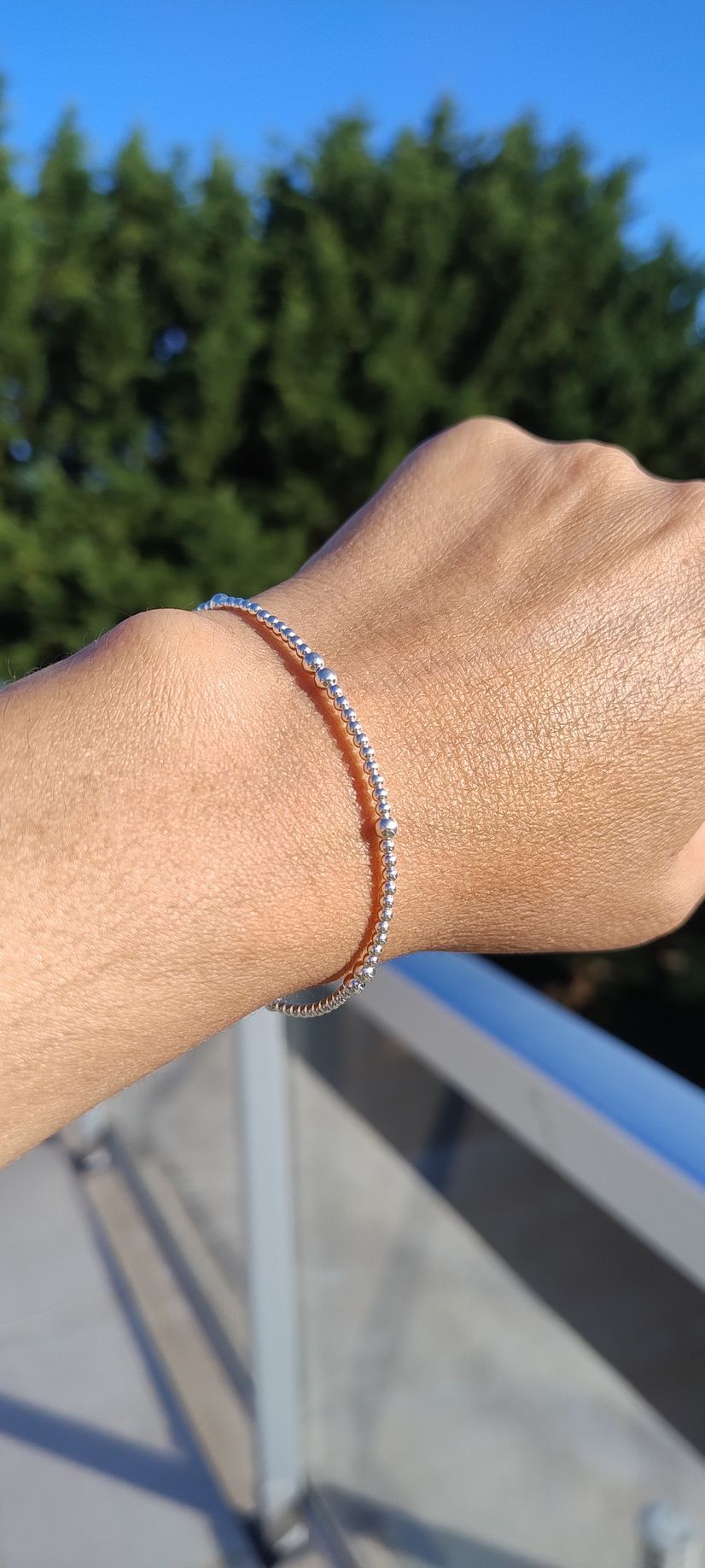 Primer plano de una mano con una fina pulsera de plata al aire libre junto a una barandilla, con árboles al fondo.