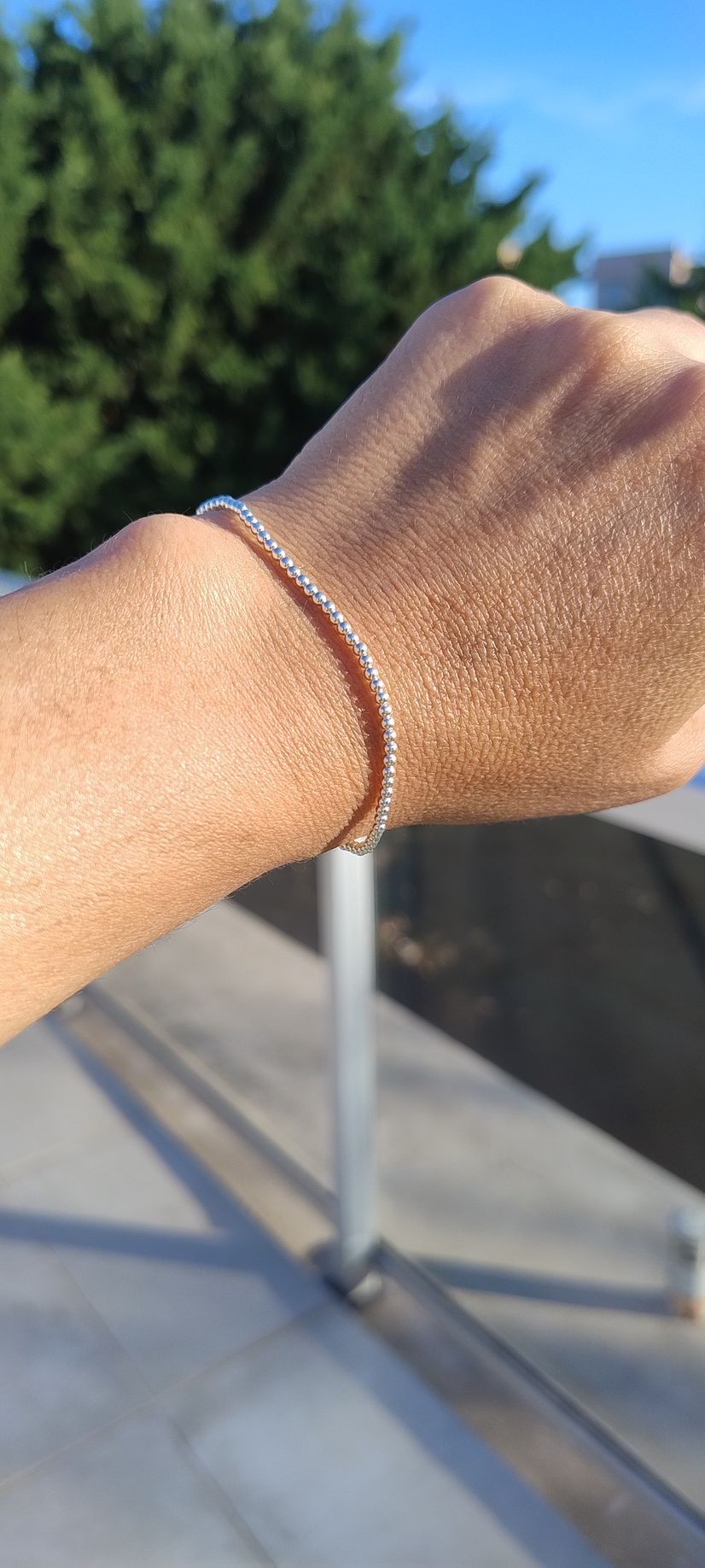 Una mano con una pulsera de plata se extendía al aire libre, bajo la luz del sol, contra una barandilla borrosa y vegetación.