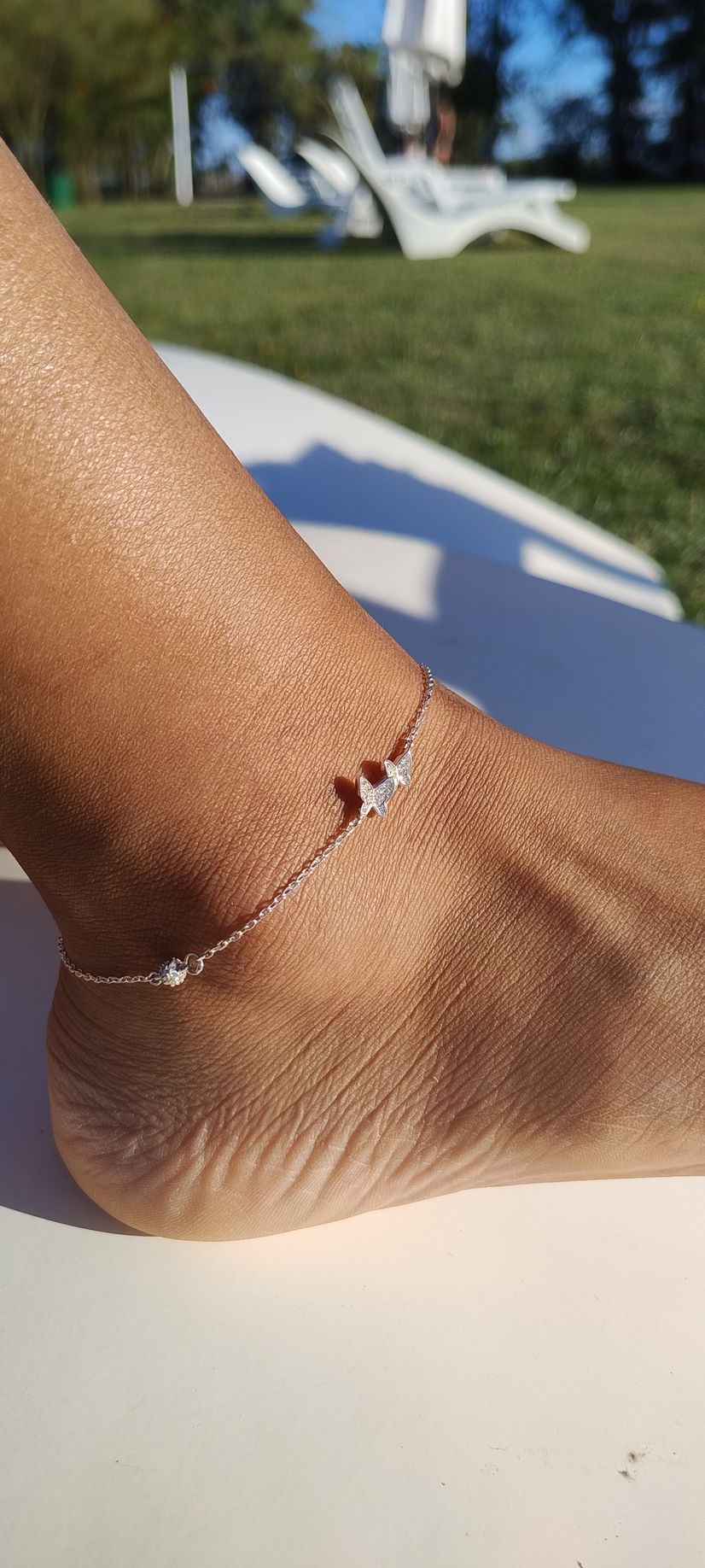 Primer plano de un tobillo con una delicada tobillera, apoyado en una tumbona al aire libre.