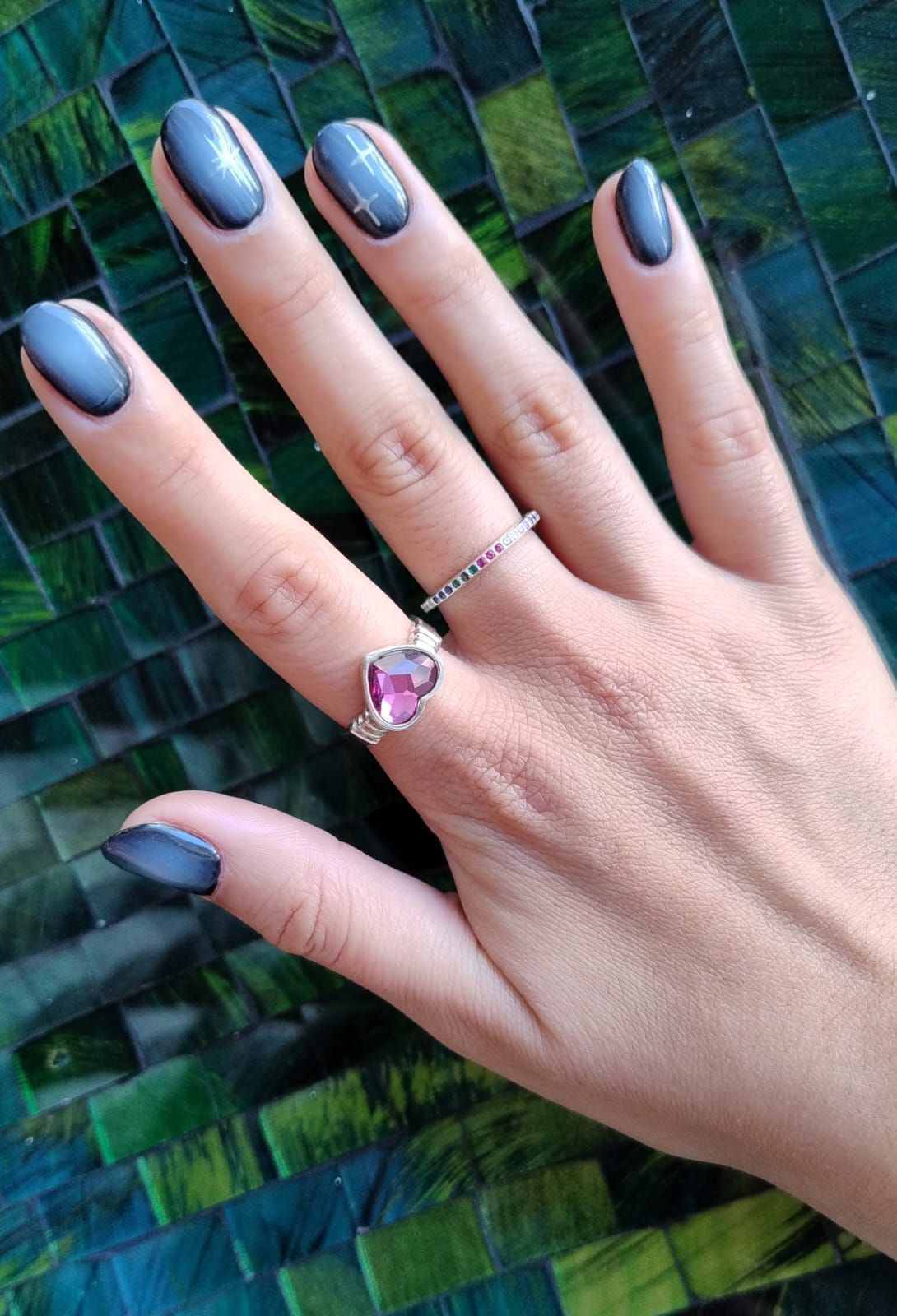 Mano con uñas azules ovaladas y dos anillos sobre fondo verde y azul. Uno de los anillos tiene un corazón rosa.