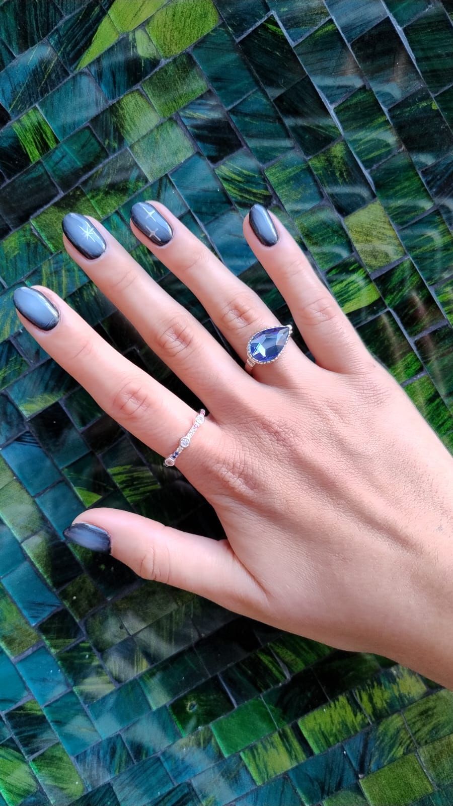 Mano con uñas azules, ribeteadas en negro, luciendo dos anillos, sobre un fondo de azulejos color verde azulado.