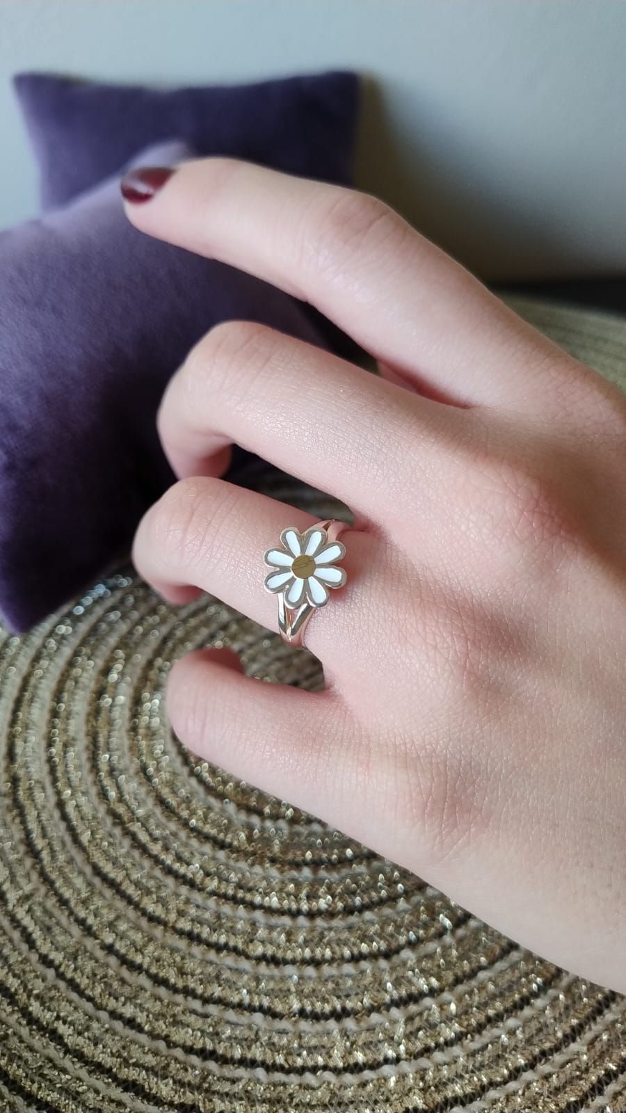 Mano con un anillo de plata con un diseño de margarita blanca, apoyada sobre una esterilla dorada;