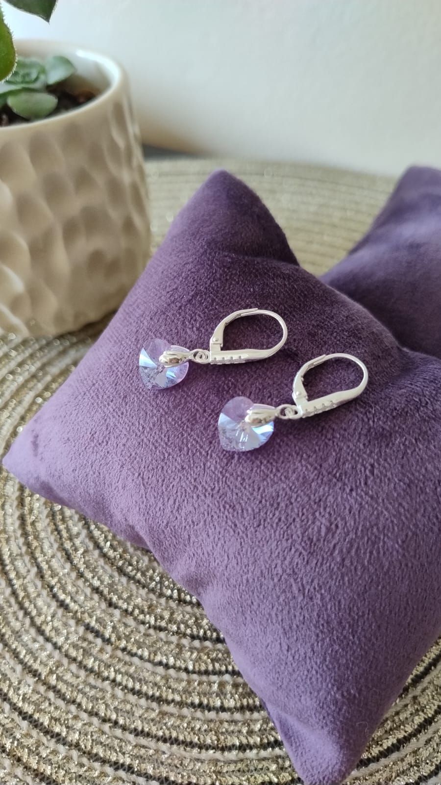 Pendientes plateados en forma de corazón con cristales azul claro, sobre un cojín de terciopelo morado.