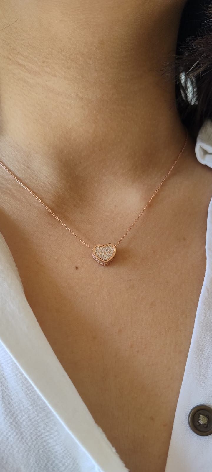 Primer plano de un cuello que luce un delicado collar de oro con un colgante en forma de corazón sobre una blusa blanca.