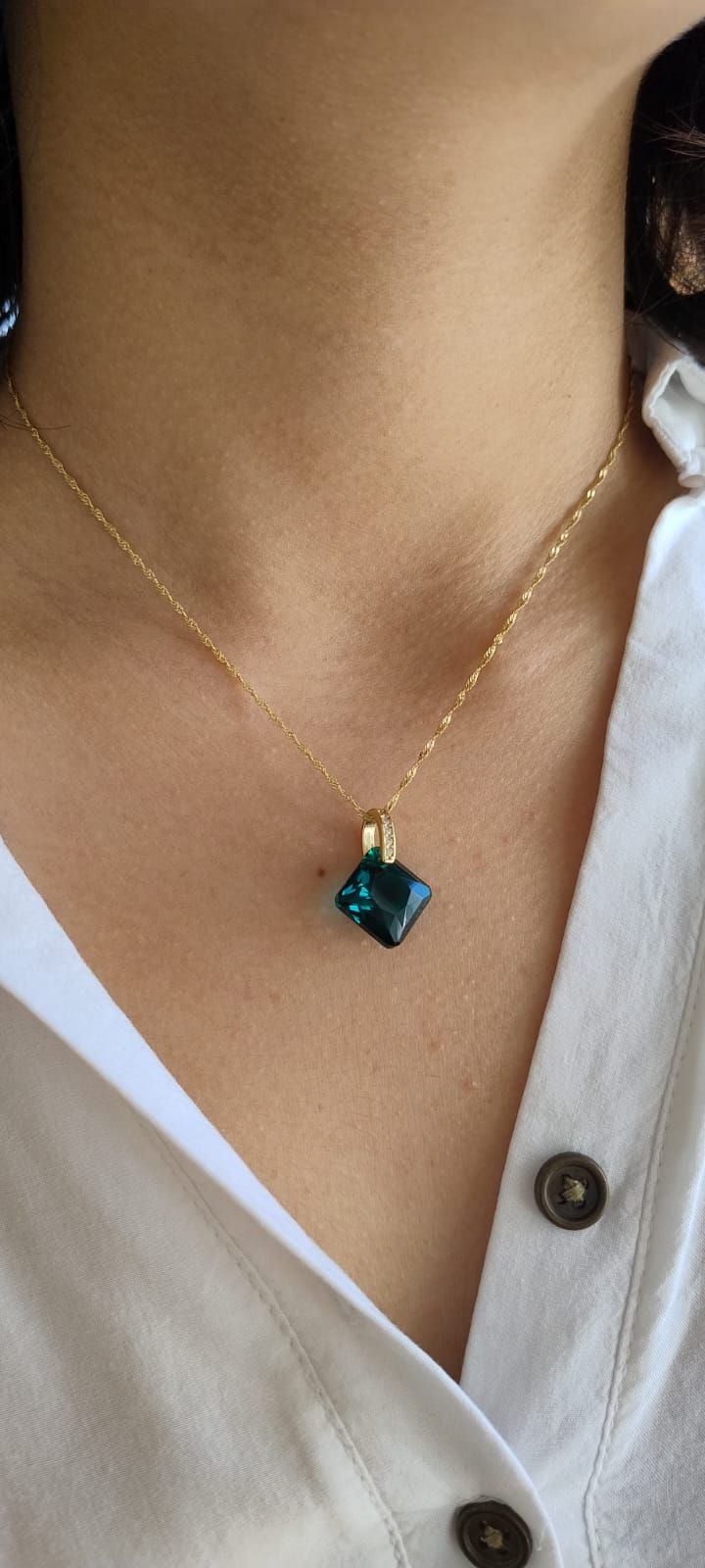 Una persona que lleva un collar de oro con un colgante cuadrado de color verde azulado y una camisa blanca.