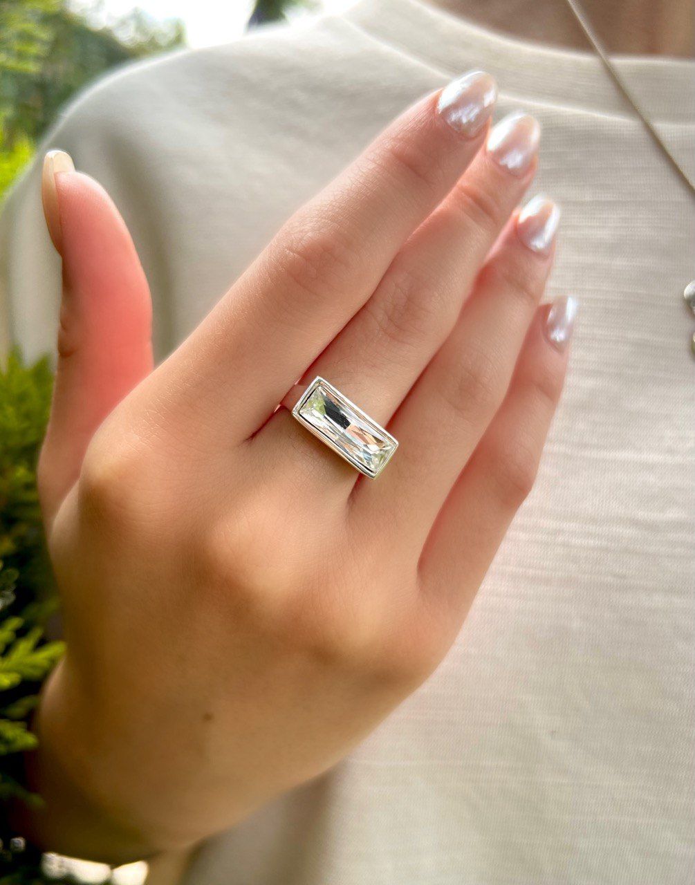 Mano sosteniendo un anillo rectangular de plata con una piedra preciosa transparente sobre un fondo de camisa clara.