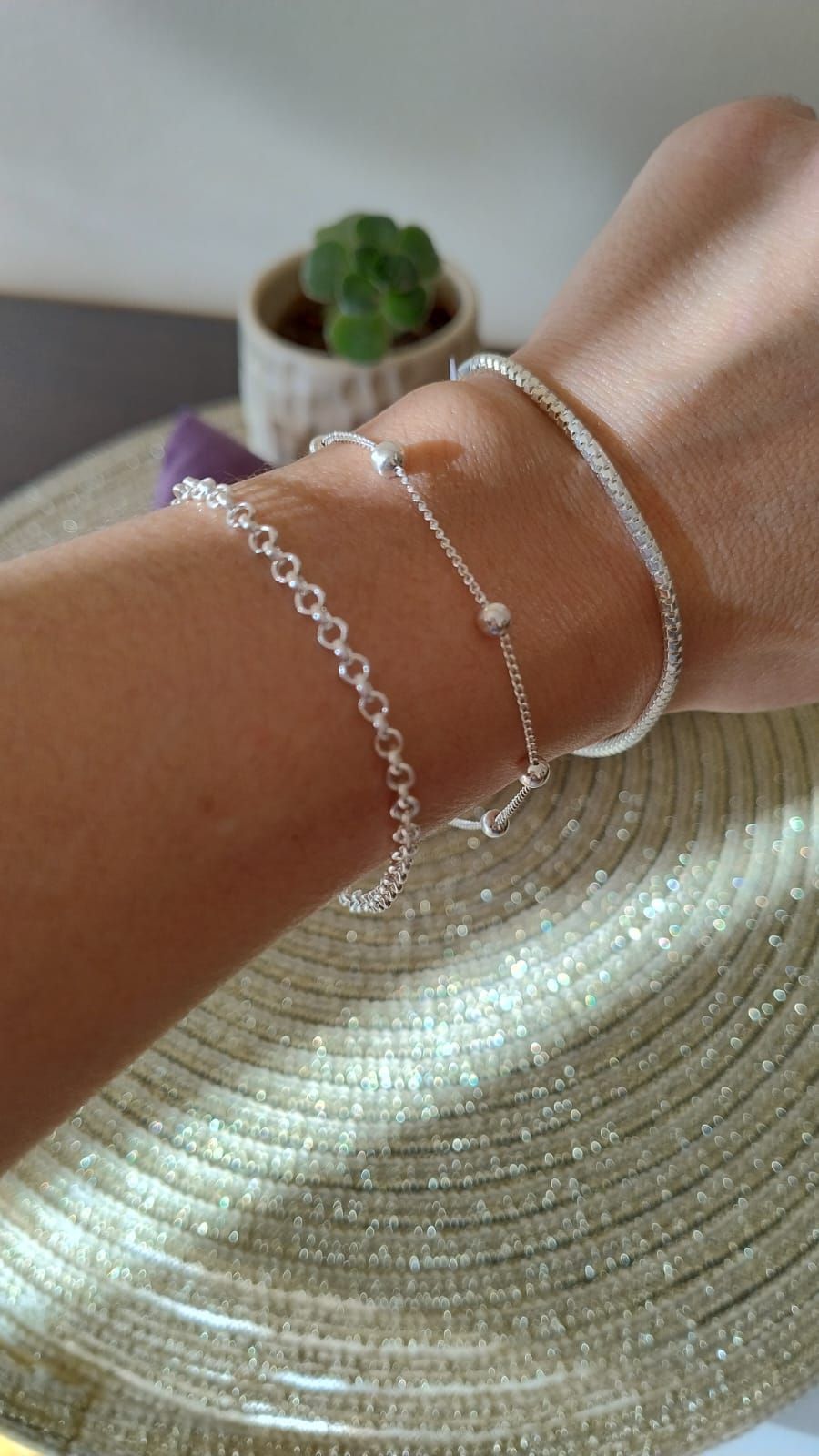 Tres delicadas pulseras de plata en una muñeca sobre un sombrero de paja tejido, con una pequeña planta suculenta al fondo.