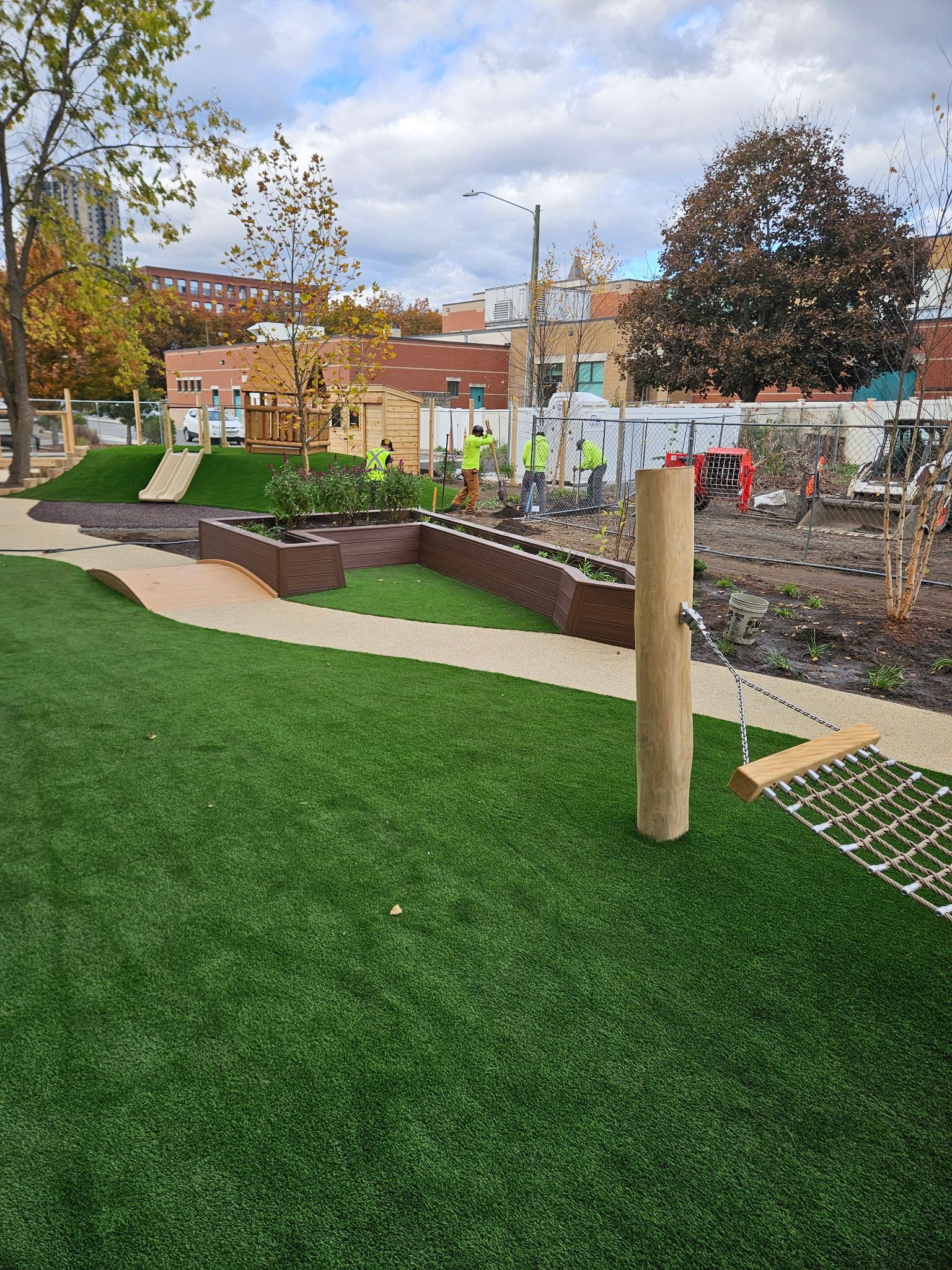 Green turf and walkway in a park with construction in the background; a hammock pole and workers are visible.