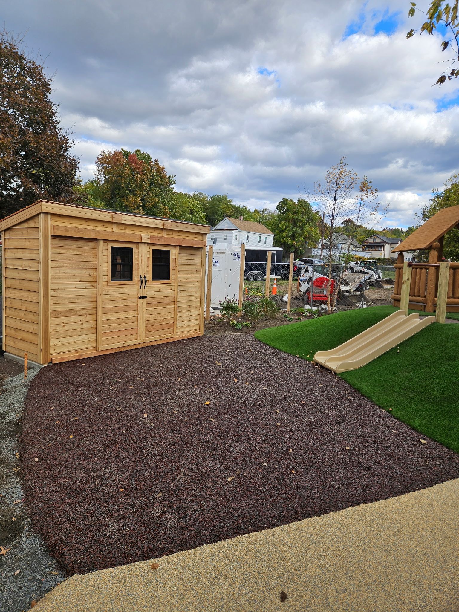 Wooden shed and playground equipment on brown mulch and green turf. Cloudy sky.