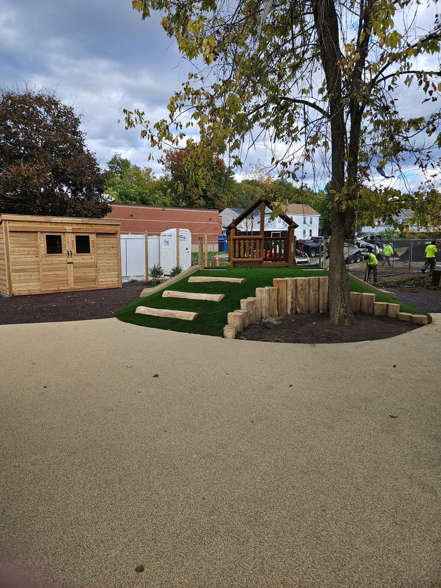 Play area with wooden structures, grass steps, and a tree, against an overcast sky.