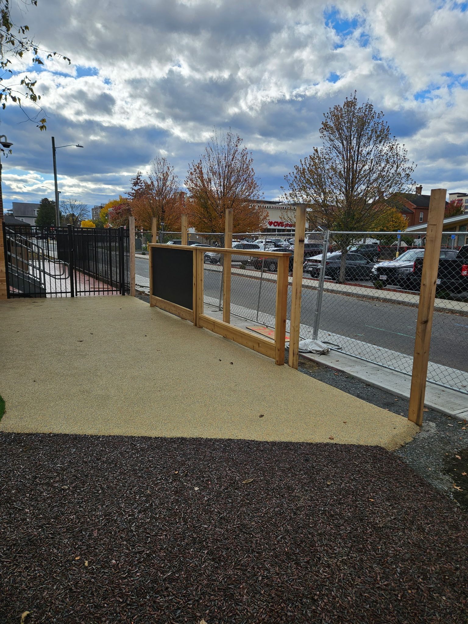 Gravel pathway with wooden fence leading to a parking area. Overcast sky, fall trees in background.