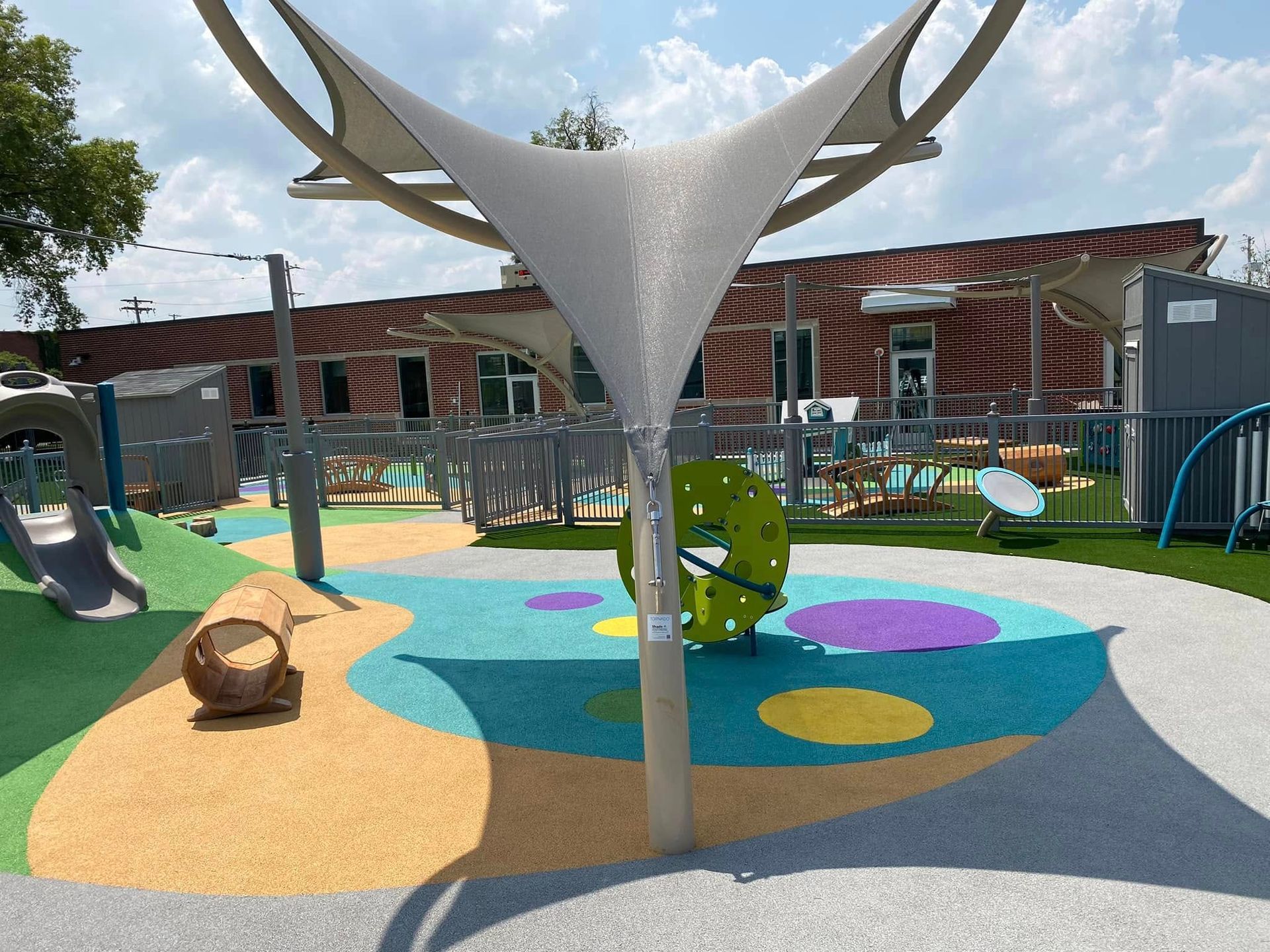 Playground with a shade structure, colorful ground, and play equipment, in front of a building.