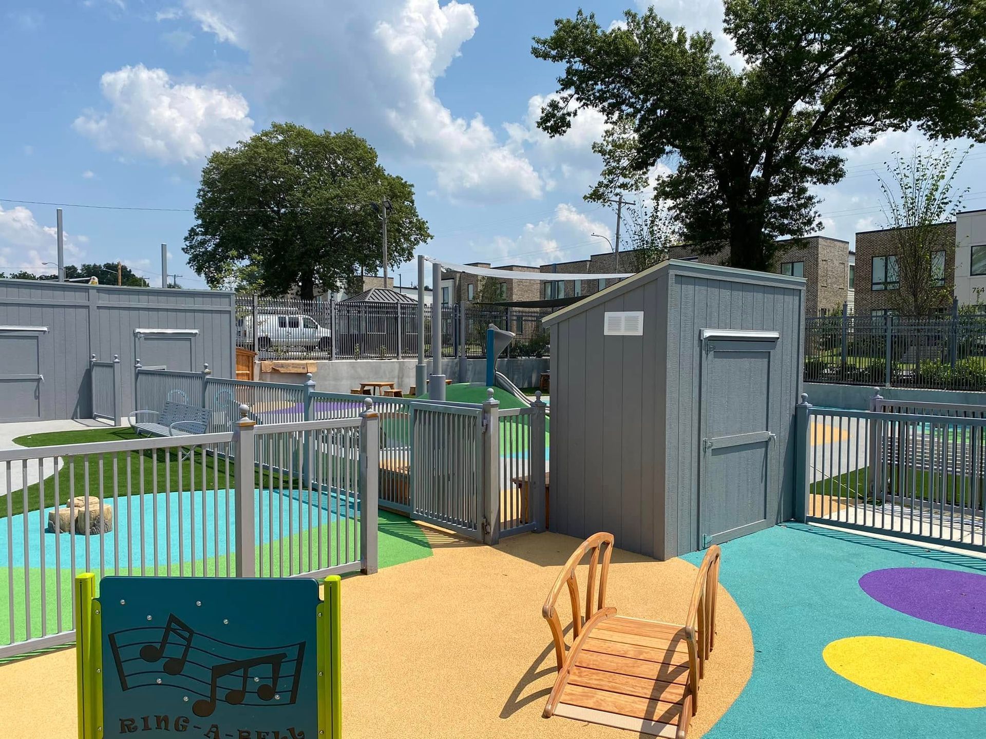 Playground with gray structures, colorful surfaces, fencing, and wooden bridge. Blue sky, green trees.