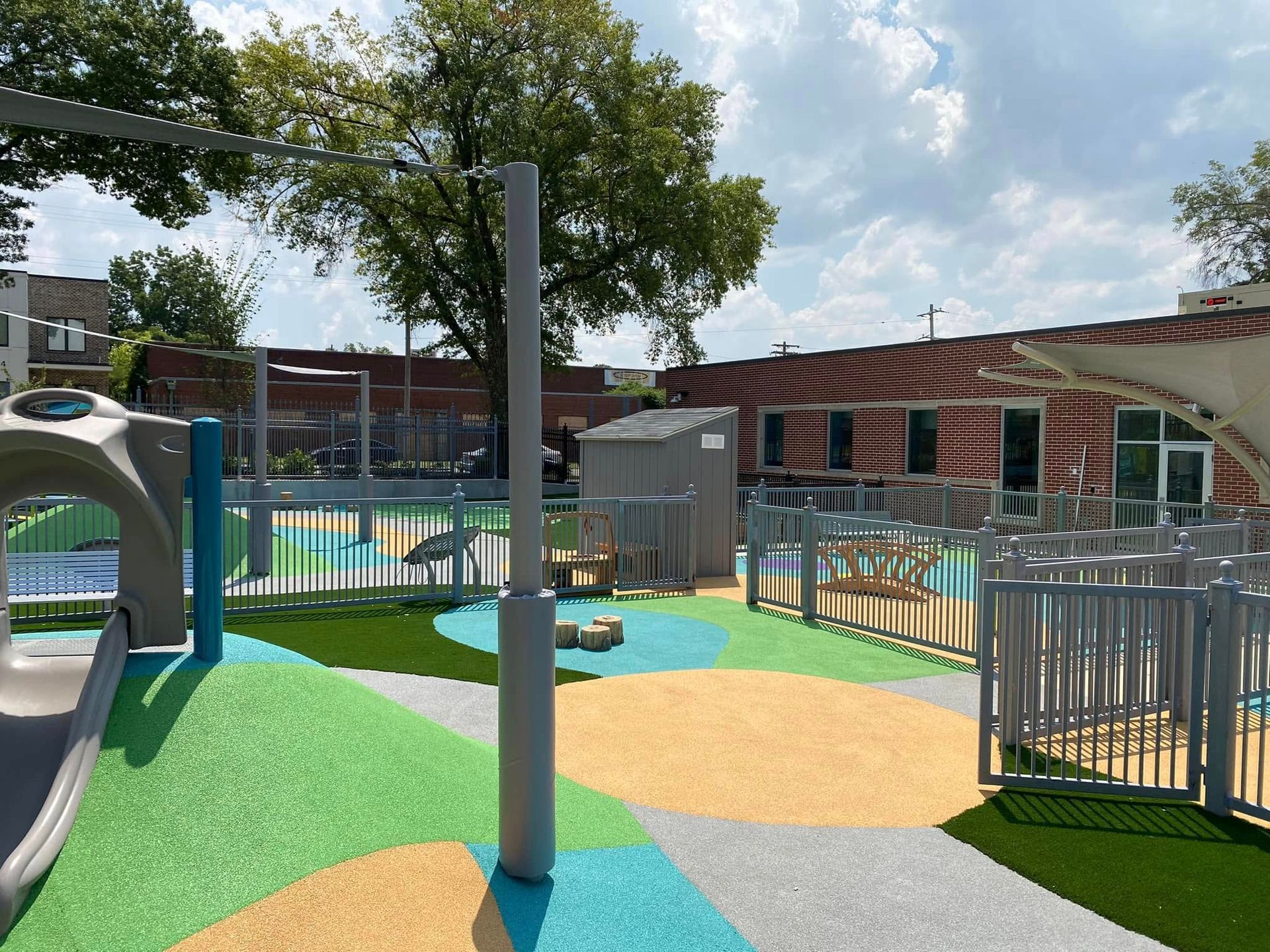 Playground with colorful turf, climbing structures, and small buildings under a cloudy sky.