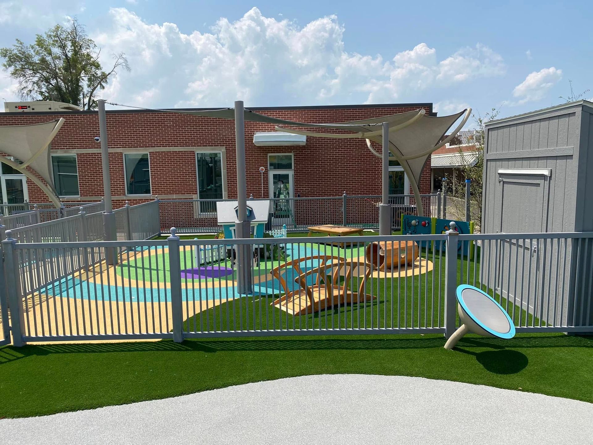 Playground with gray fence, artificial turf, and colorful play area, with shade sails and a building in the background.