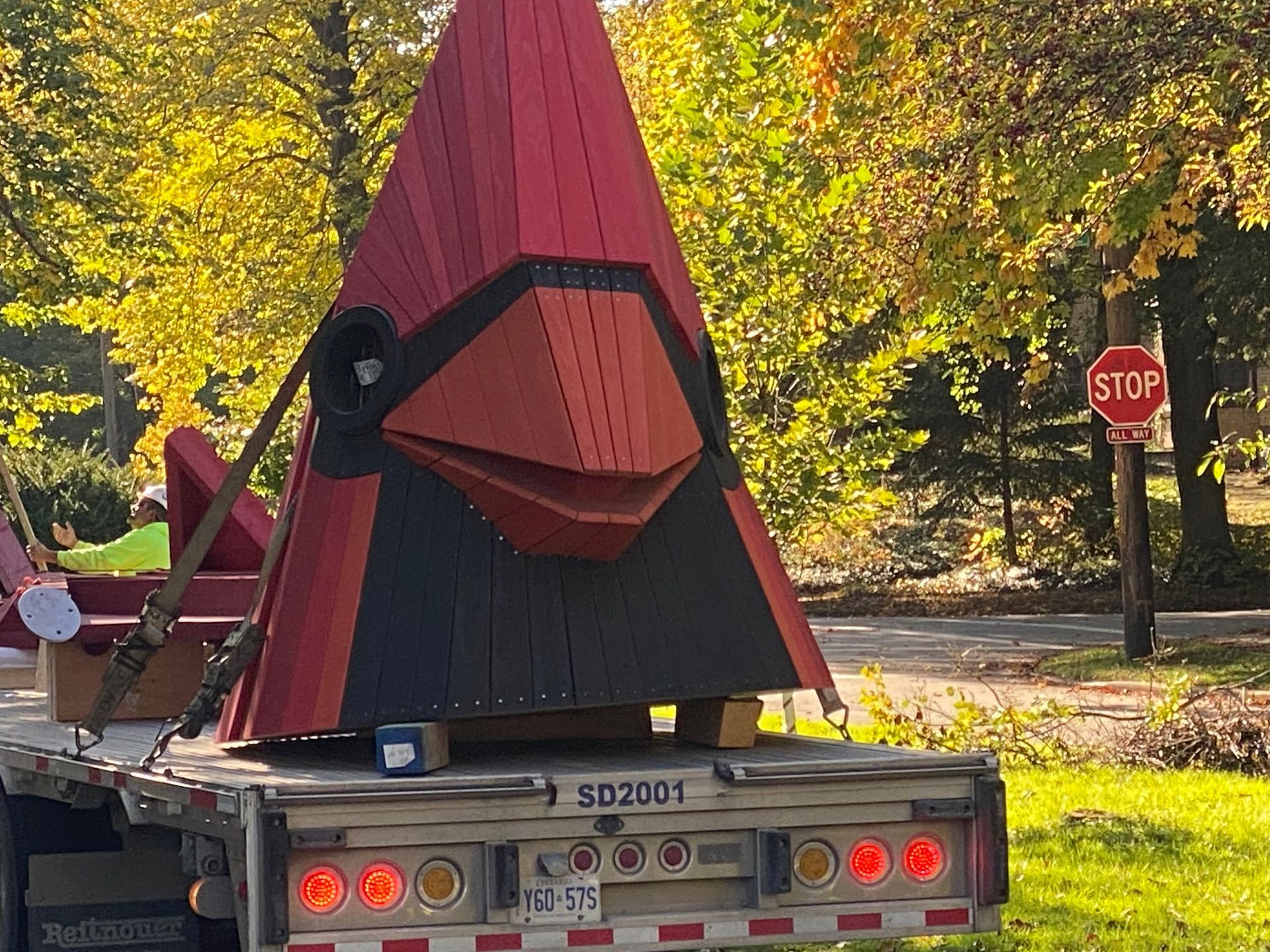 Large, red and black cardinal sculpture on a flatbed truck, near a stop sign, with fall foliage in the background.
