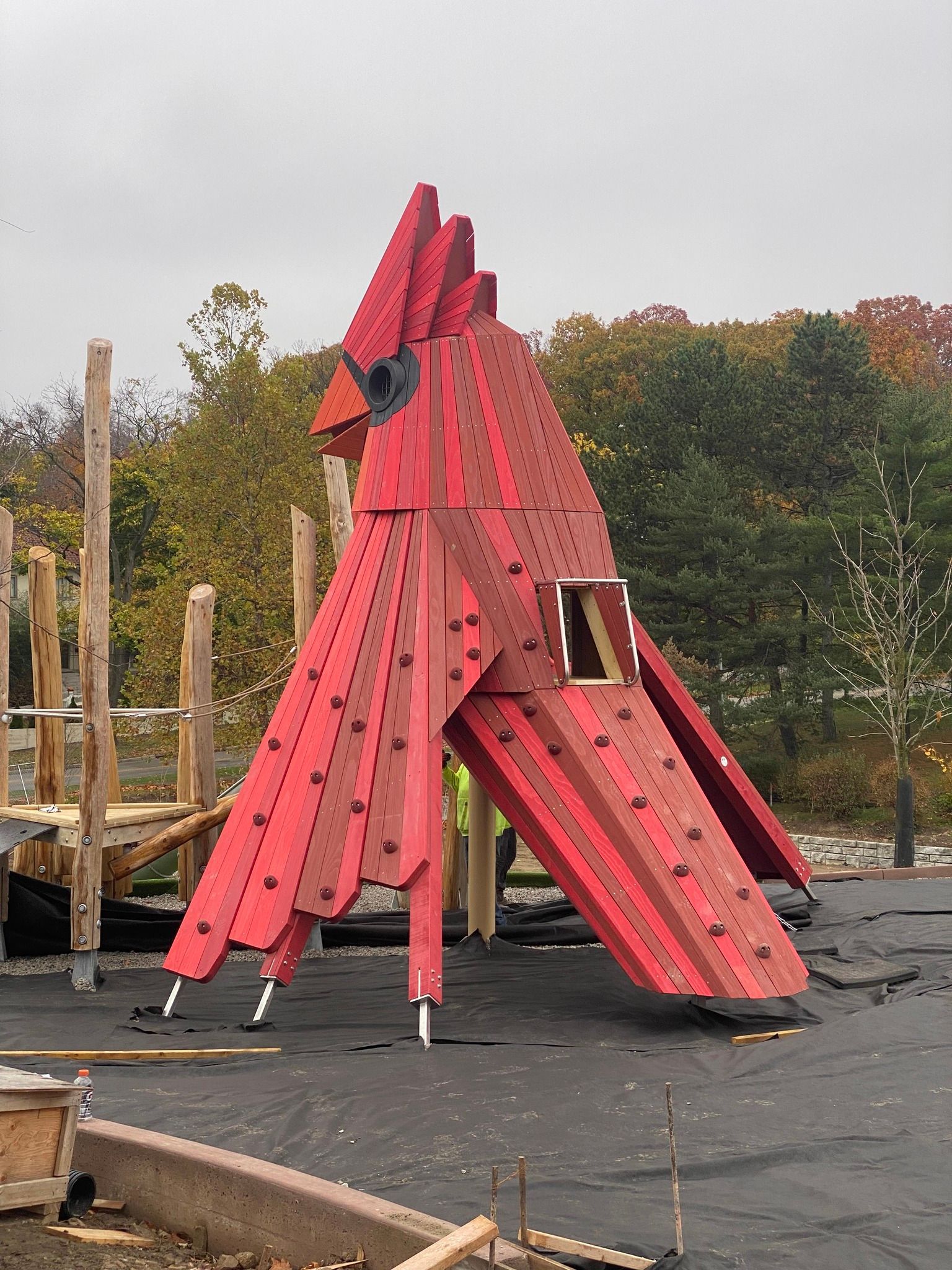 Red cardinal-shaped playground climbing structure with a window, on asphalt, with trees in the background.
