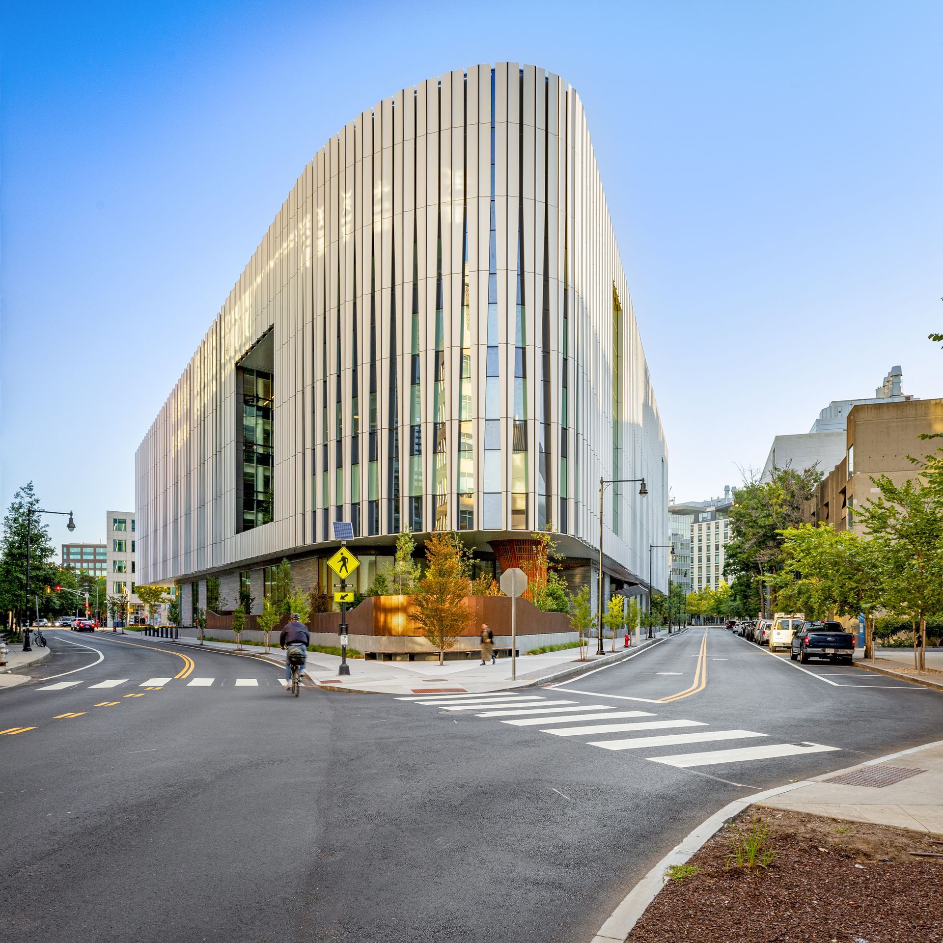 Modern, curved building with vertical white fins, at a street intersection. A cyclist rides in the foreground.