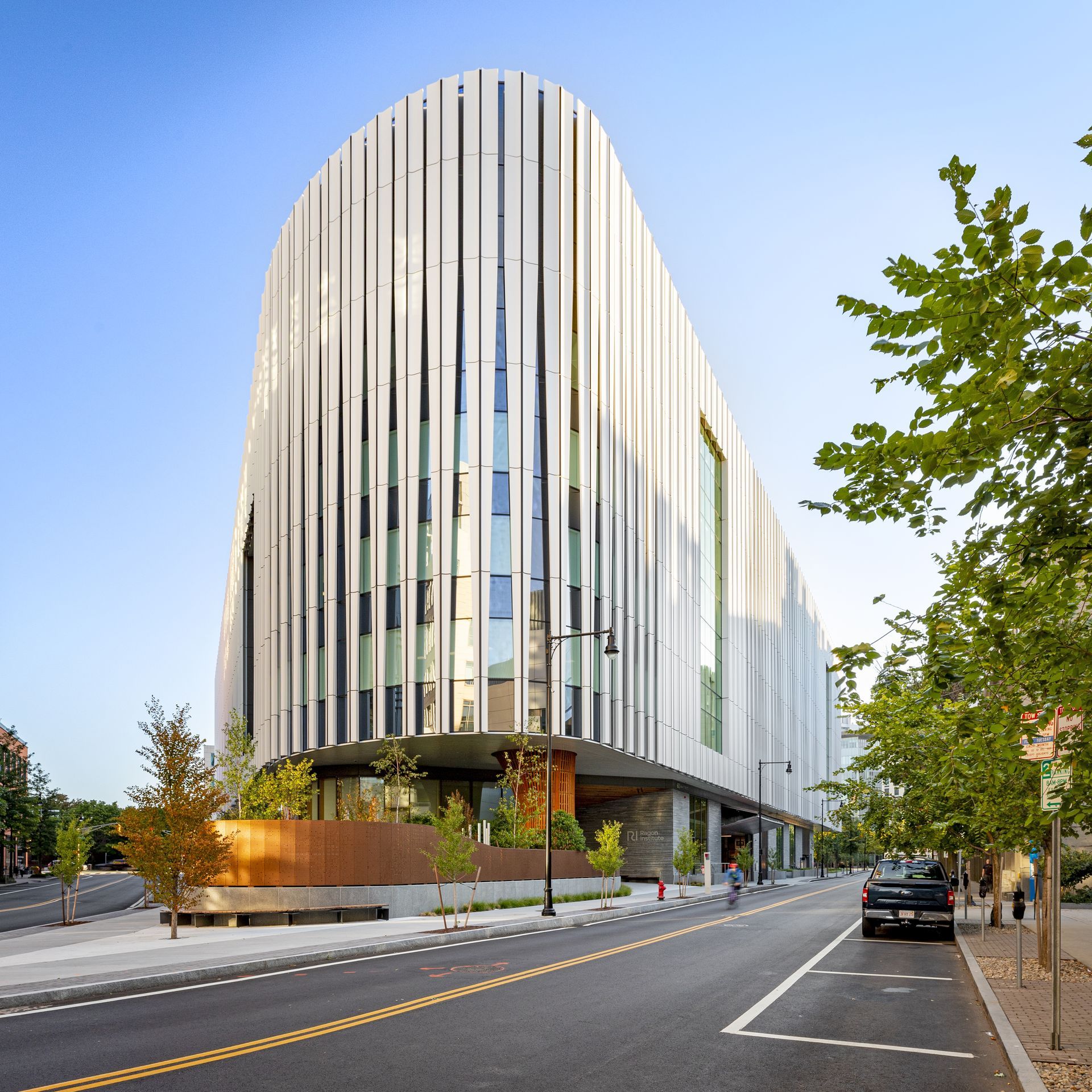 Modern, white, vertically-striped building with curved facade; trees and a street in the foreground.