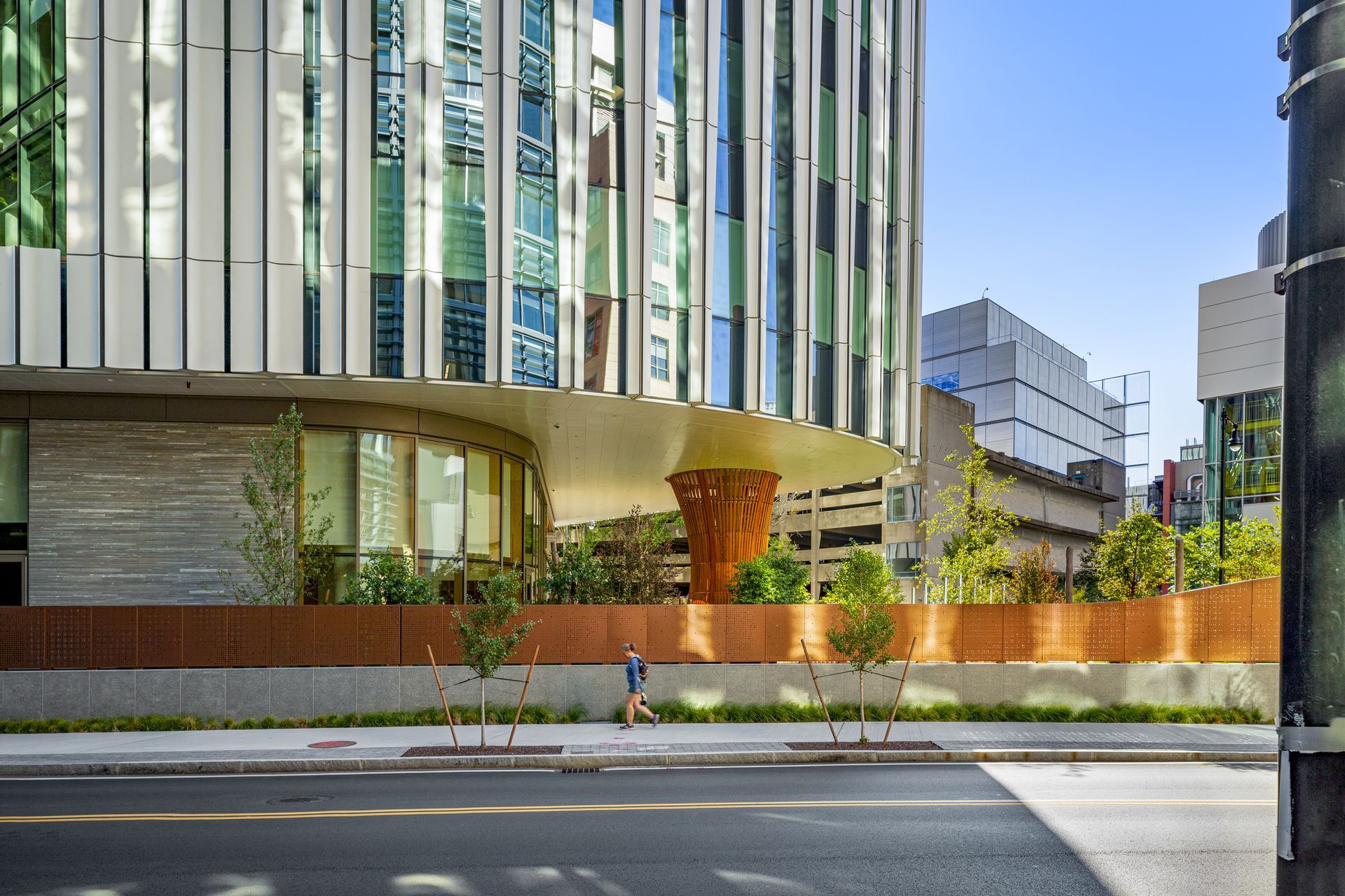 Modern building with curved base and vertical stripes, landscaping with pedestrian on sidewalk.