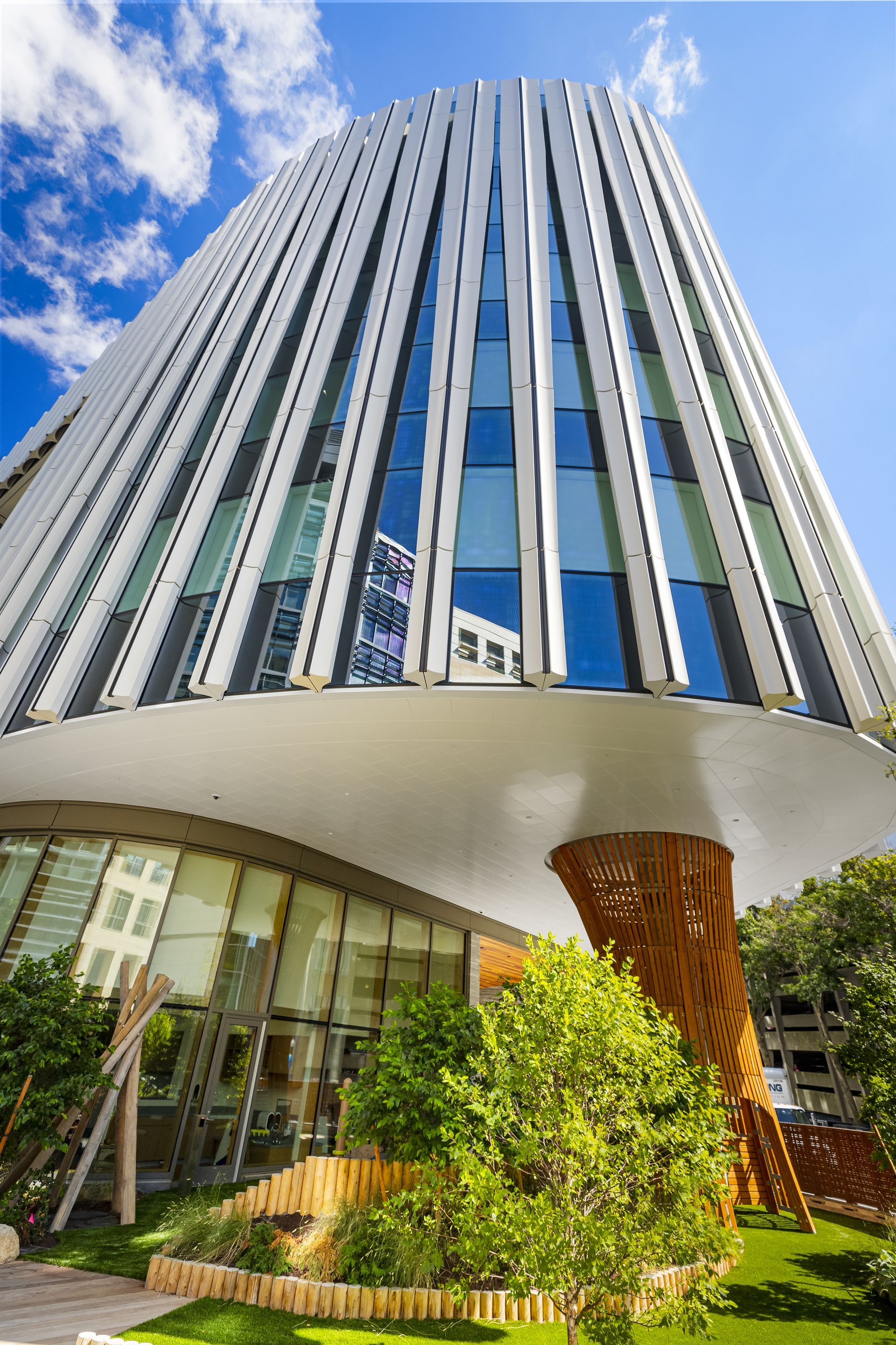Modern building with white vertical panels, glass windows, and a curved base; trees in the foreground against a blue sky.