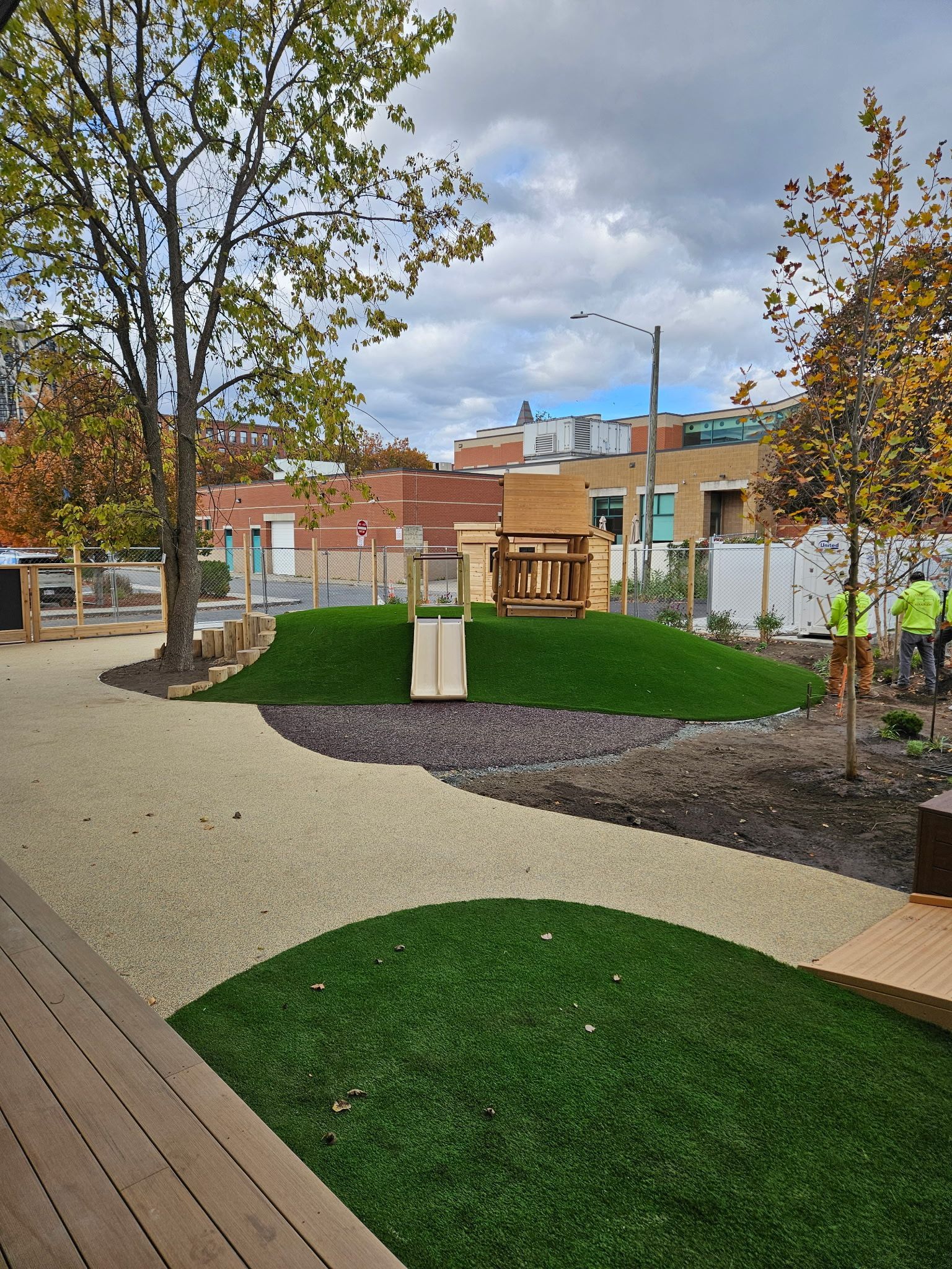 Playground with a small hill, slide, and wooden structure; gravel and artificial turf ground.
