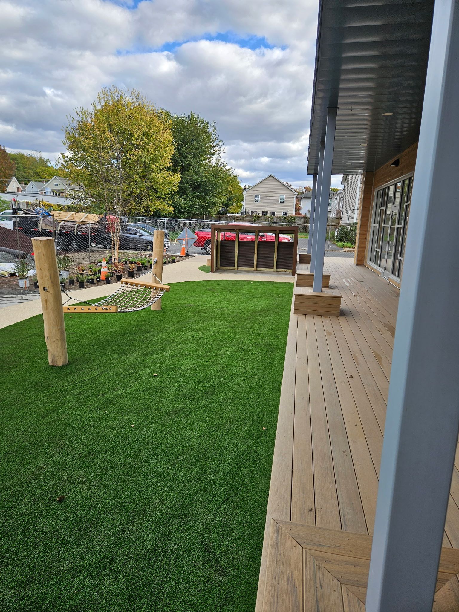 Artificial grass lawn next to a wooden deck. Trees and a small bar are in the background. Cloudy sky.