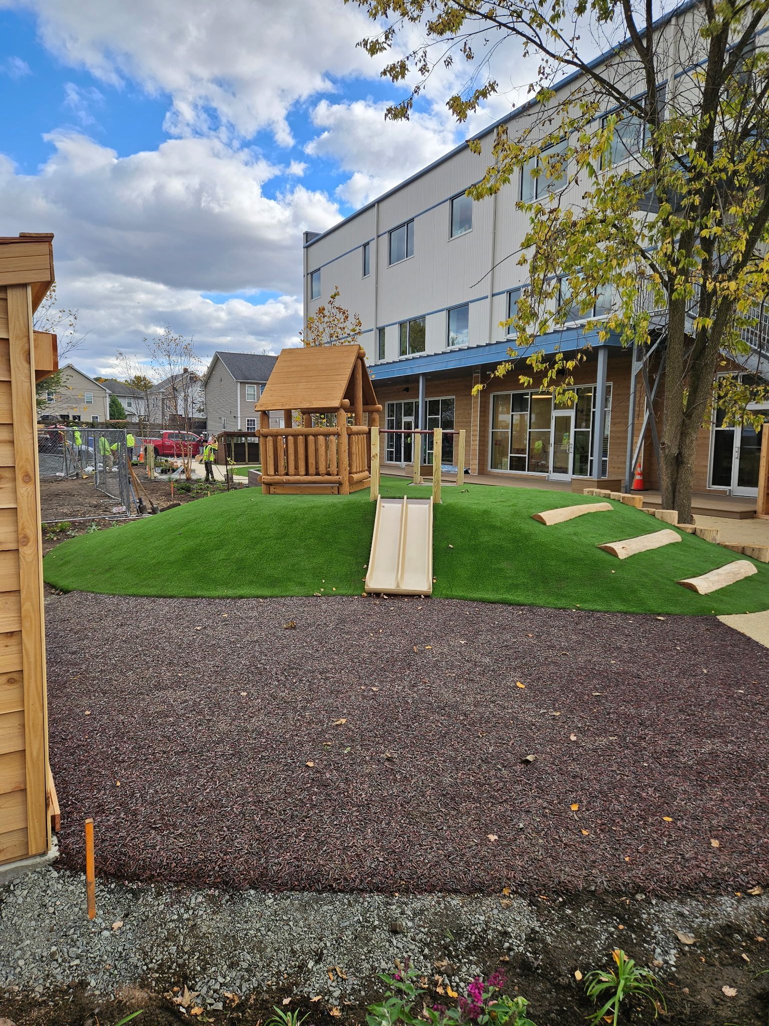 Playground with wooden structures on a grassy hill, adjacent to a building. Ground cover is dark brown.