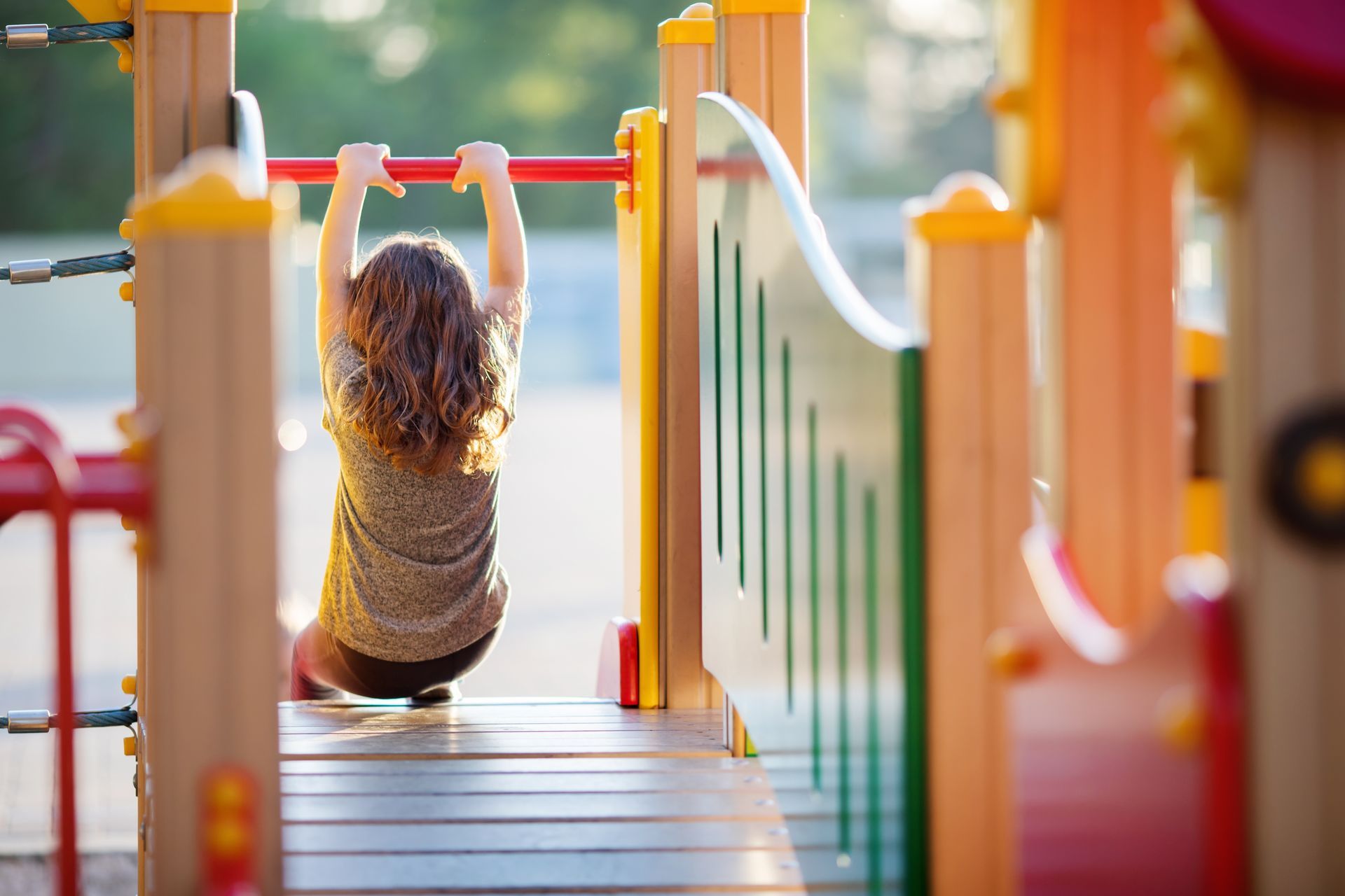 Child hanging from a horizontal bar on a colorful playground, viewed from behind.