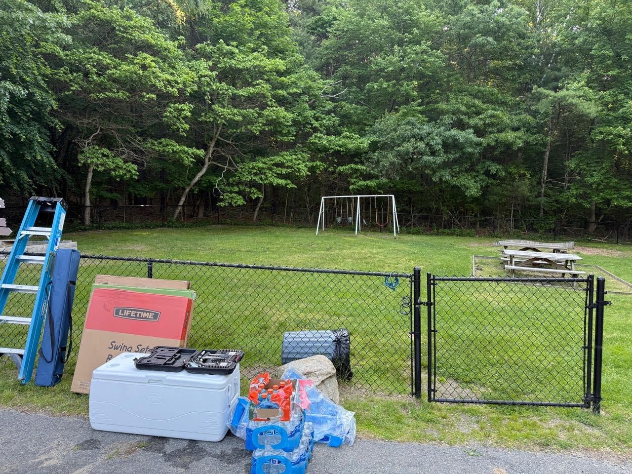 Lawn with a chain-link fence, soccer goal, picnic tables, and various items like a ladder and cooler. Green trees in the background.