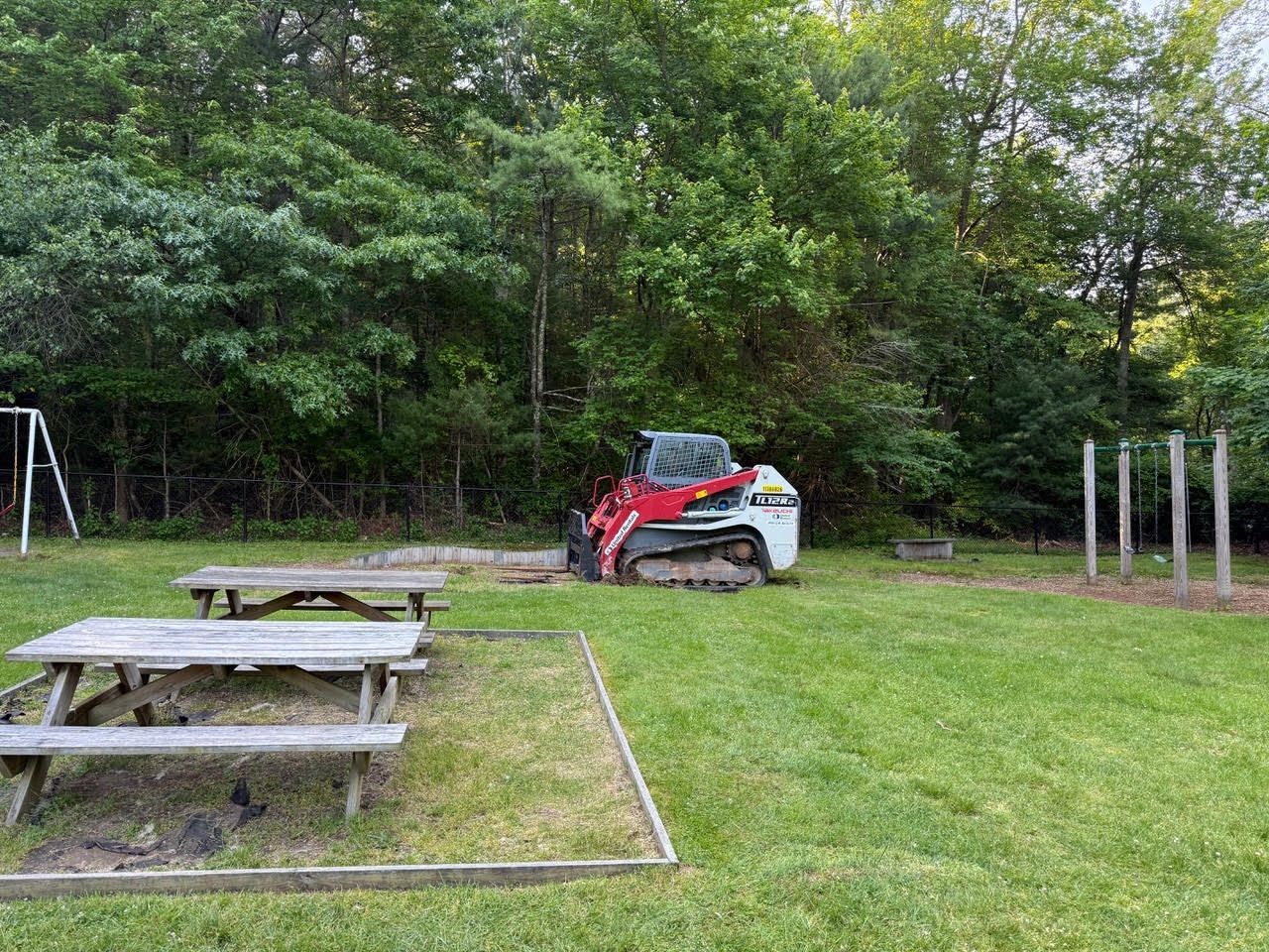 A small Bobcat excavator on grass near picnic tables and wooded area.
