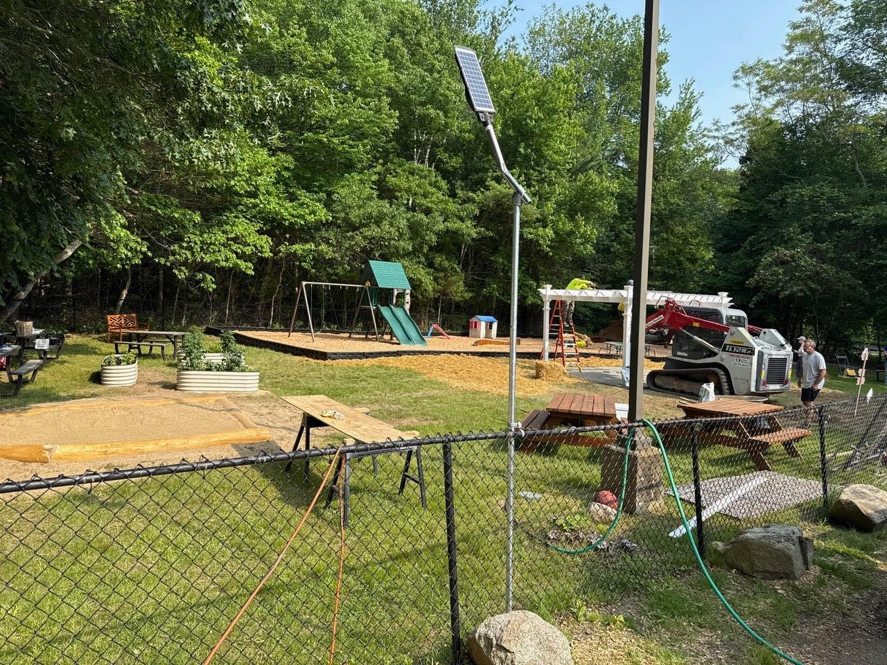 A fenced playground with mulch, swings, a slide, picnic tables, and a solar-powered light. A small excavator is present.