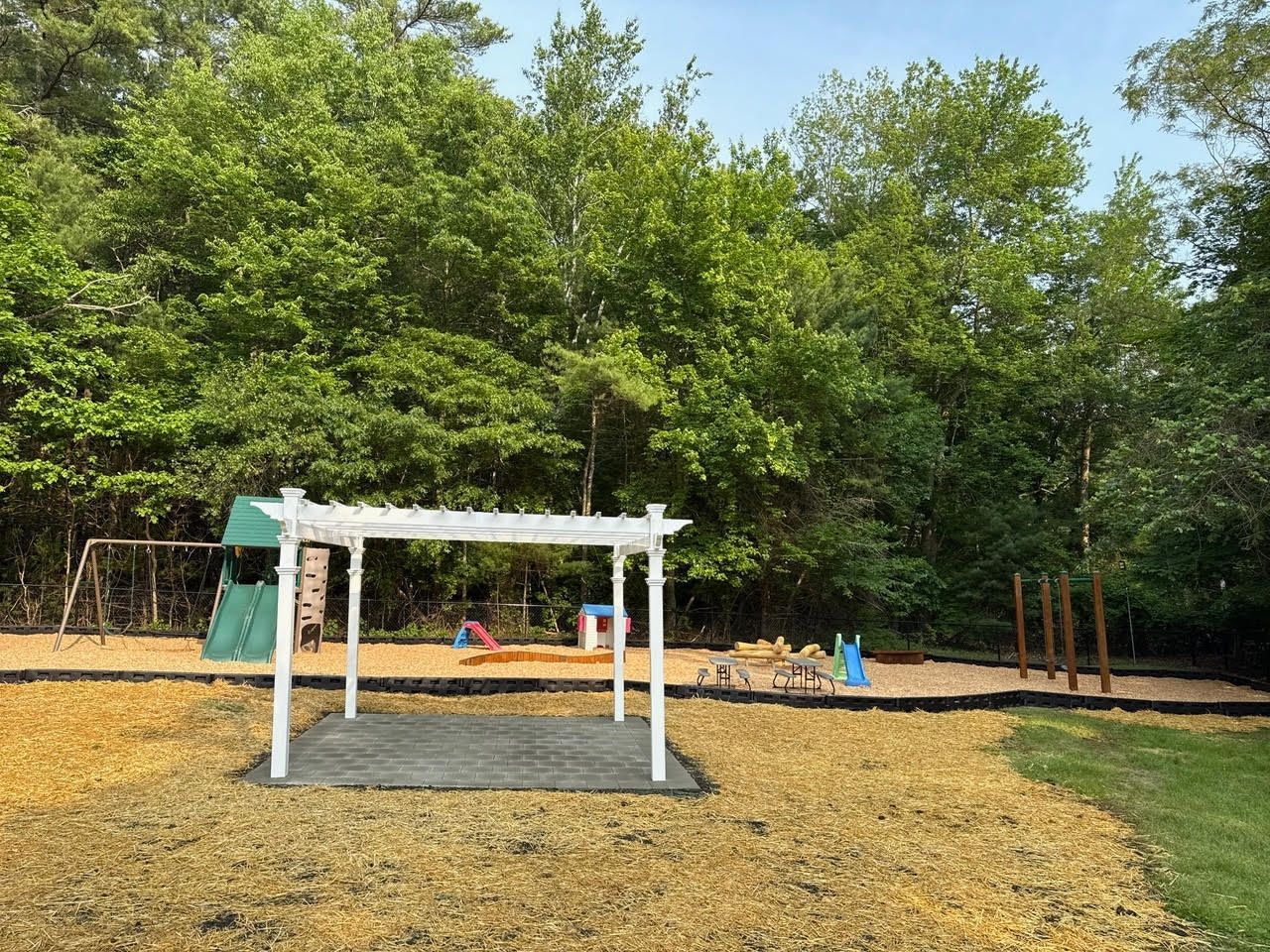 Playground with white pergola and swings in a wooded area, surrounded by wood chips.