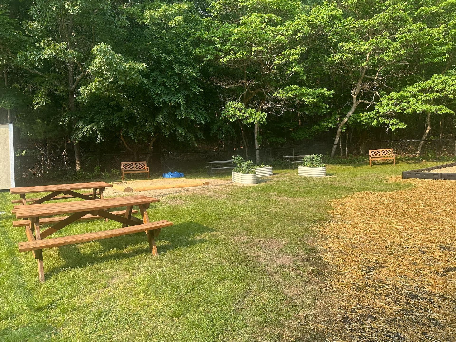Picnic tables, benches, and raised garden beds in a grassy clearing surrounded by trees.