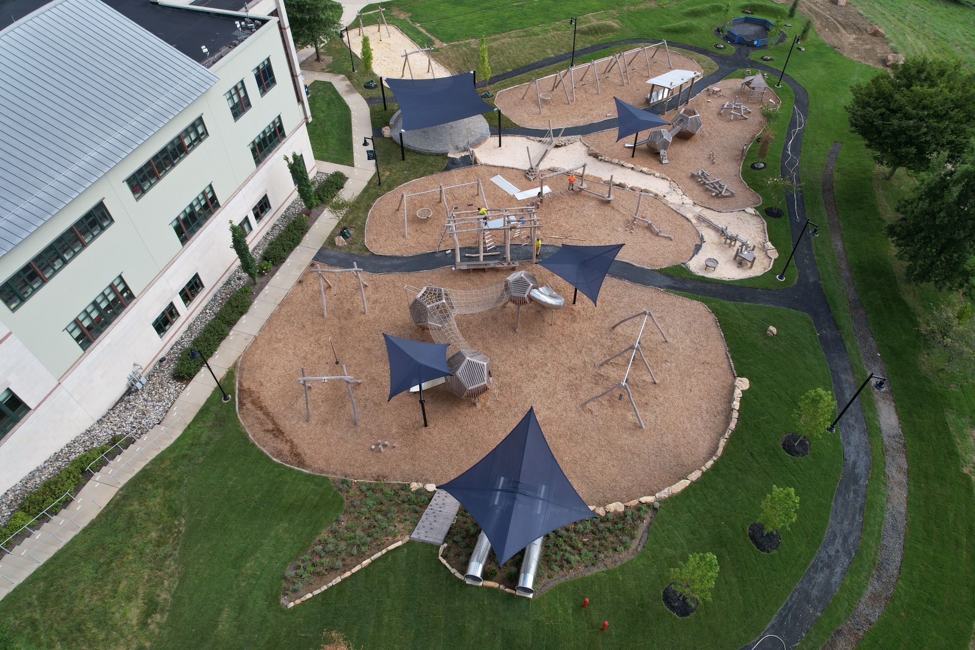 Aerial view of a playground with wood chips, climbing structures, and shaded areas. Adjacent to a building and green grass.