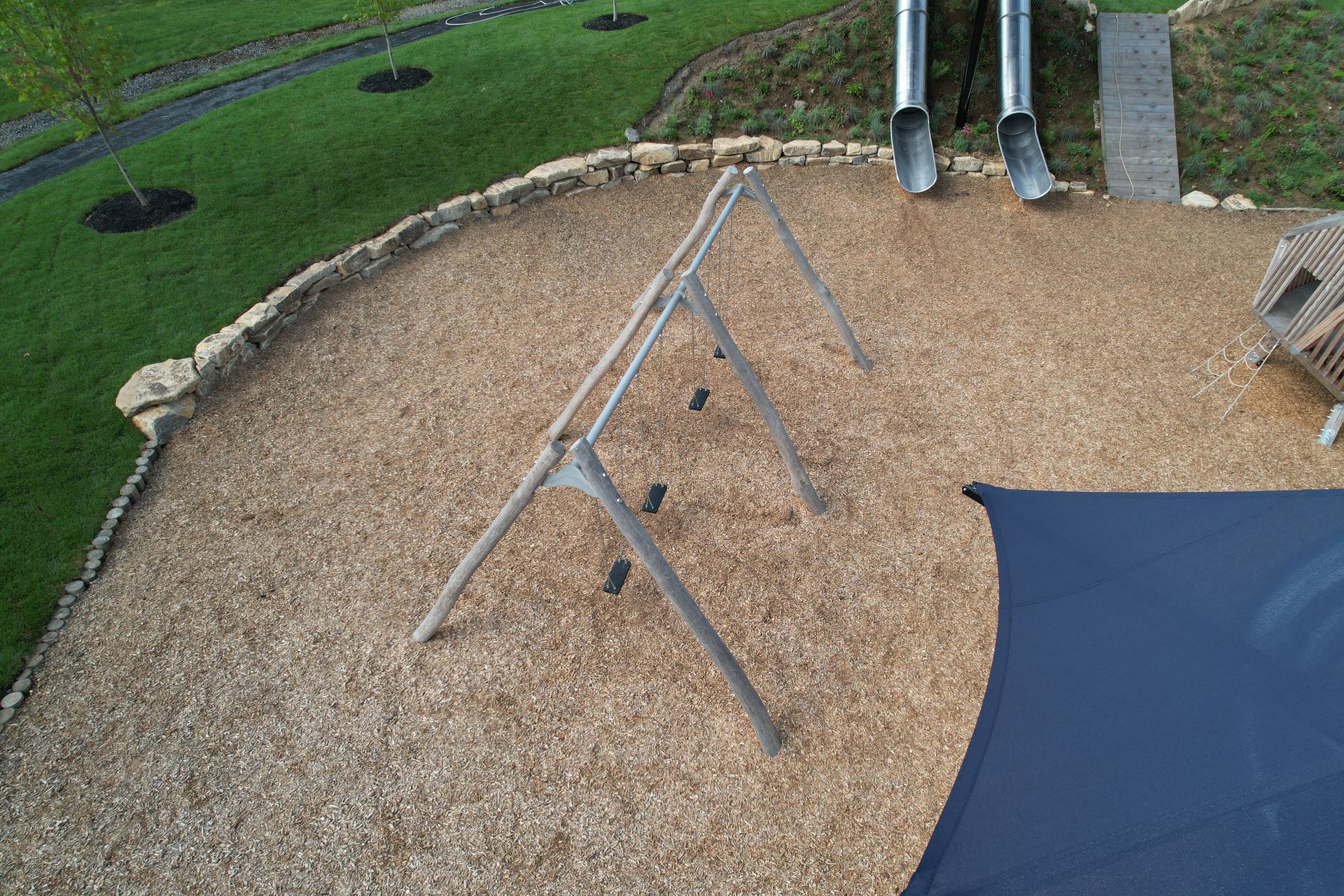 Overhead view of a playground with swing set, slides, wood chip ground cover, and shade sail.