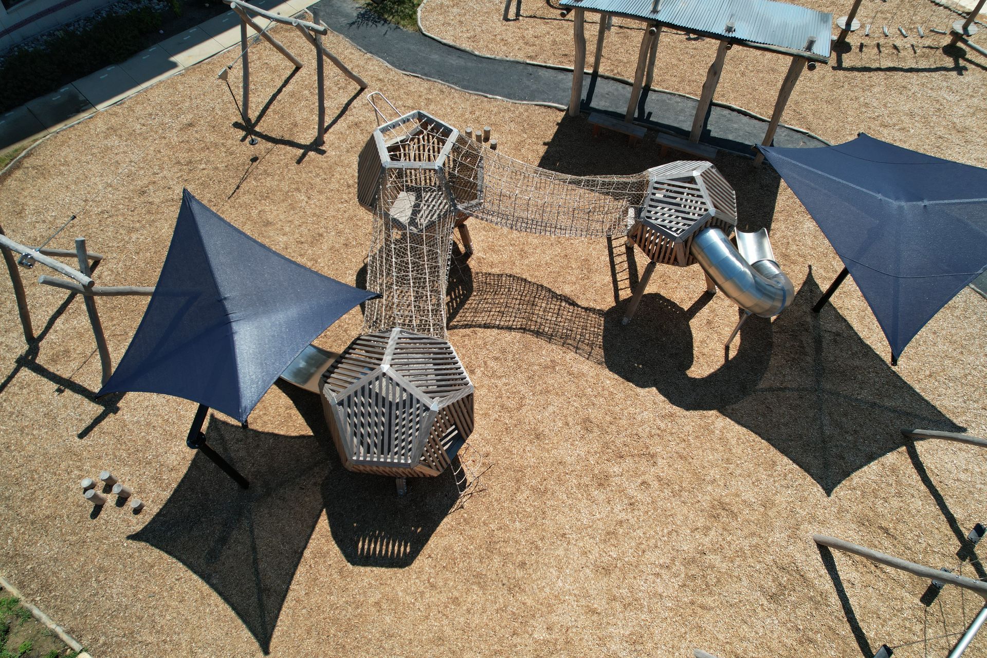 Playground with wooden structures, metal slides, and shade sails, surrounded by wood chips.