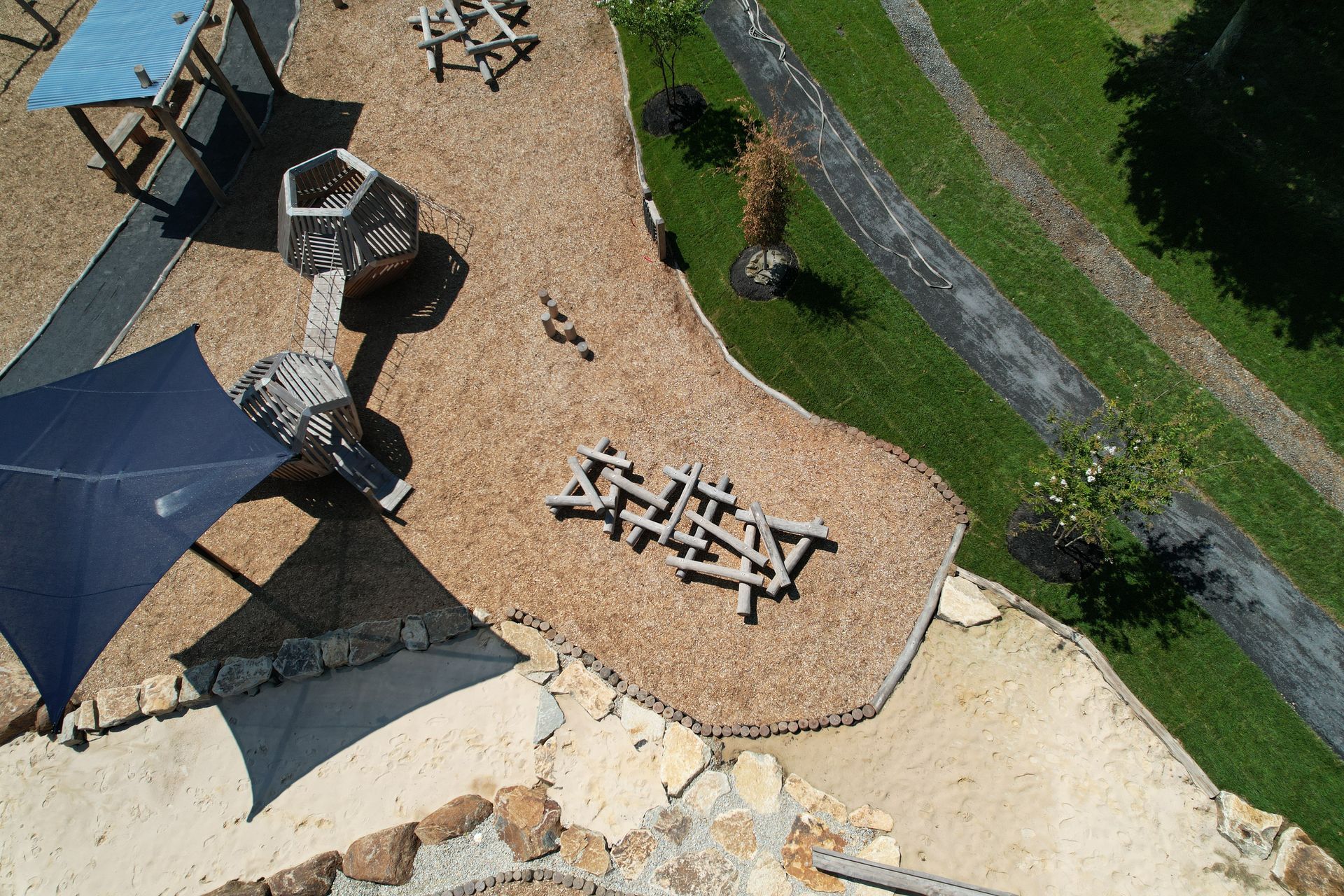 Overhead view of a playground with wood chips, wooden structures, shade sails, and a grassy area.