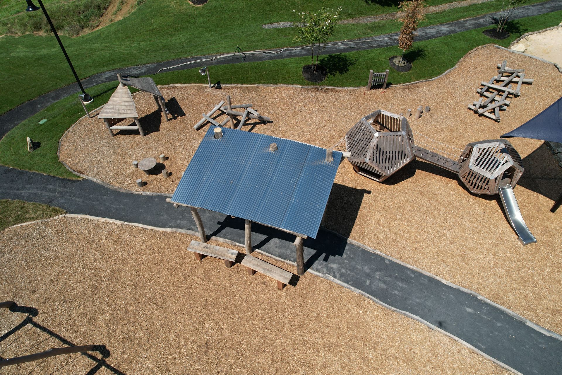 Playground with wooden structures, including a shelter, climbing structure, and slide, on a bed of wood chips.