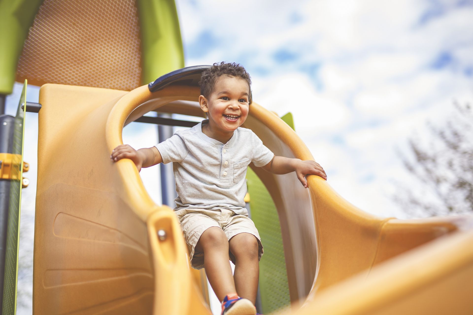 A child sits at the top of a yellow slide on a playground.