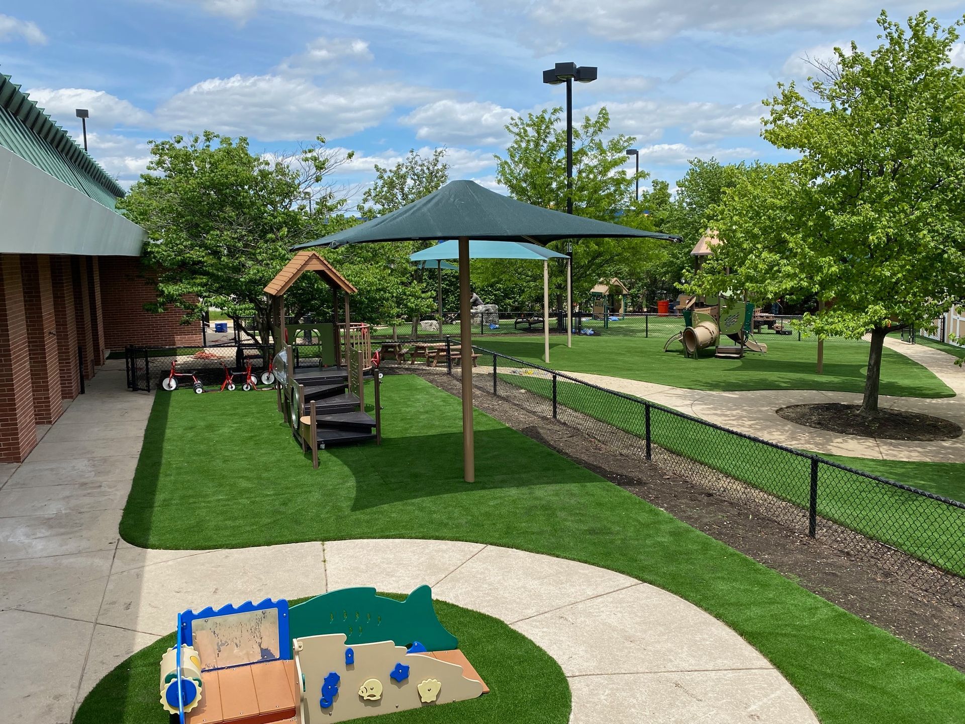 An outdoor playground featuring green artificial turf, a shaded play structure, and a colorful sand table.