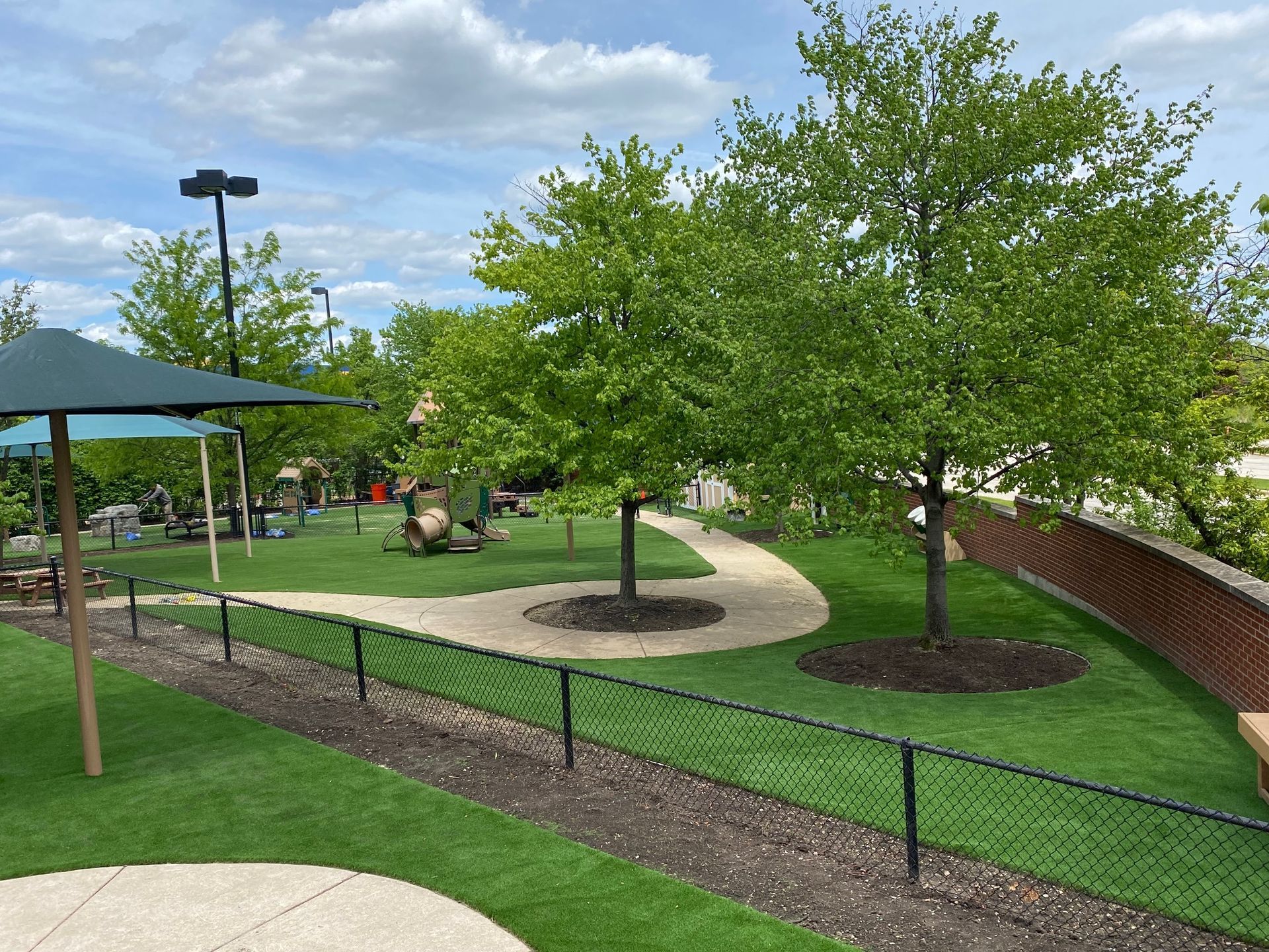 A sunlit park with green trees, a winding stone path, a chain-link fence, and a blue shade umbrella over a lawn.