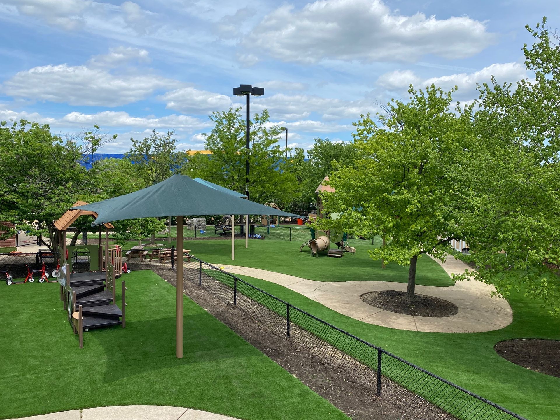 A green umbrella over a playground area with a winding stone path, manicured grass, and trees under a blue, cloudy sky.