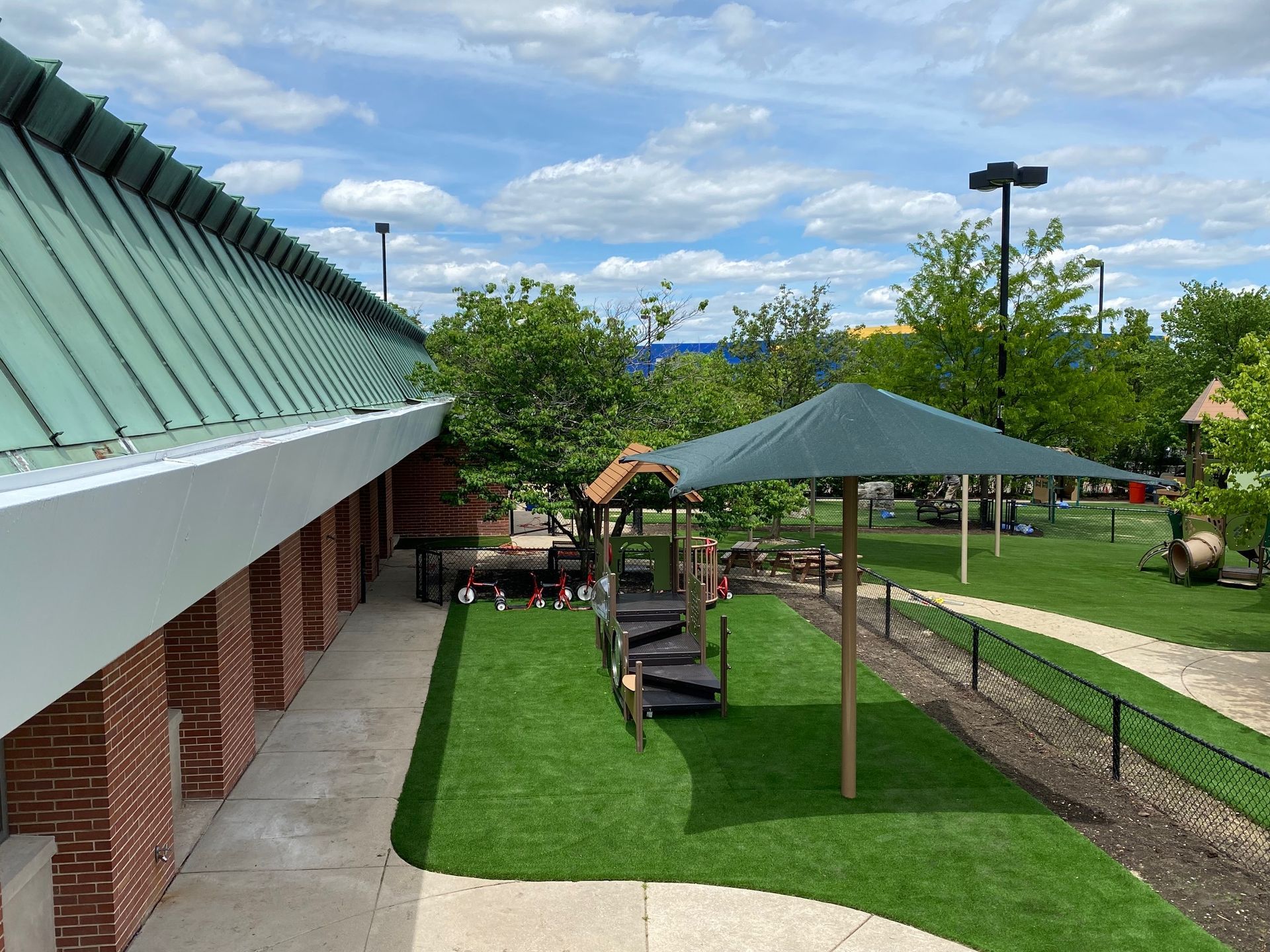 Outdoor playground with a green shade canopy, picnic tables, and a brick-walled building under a bright, cloudy sky.
